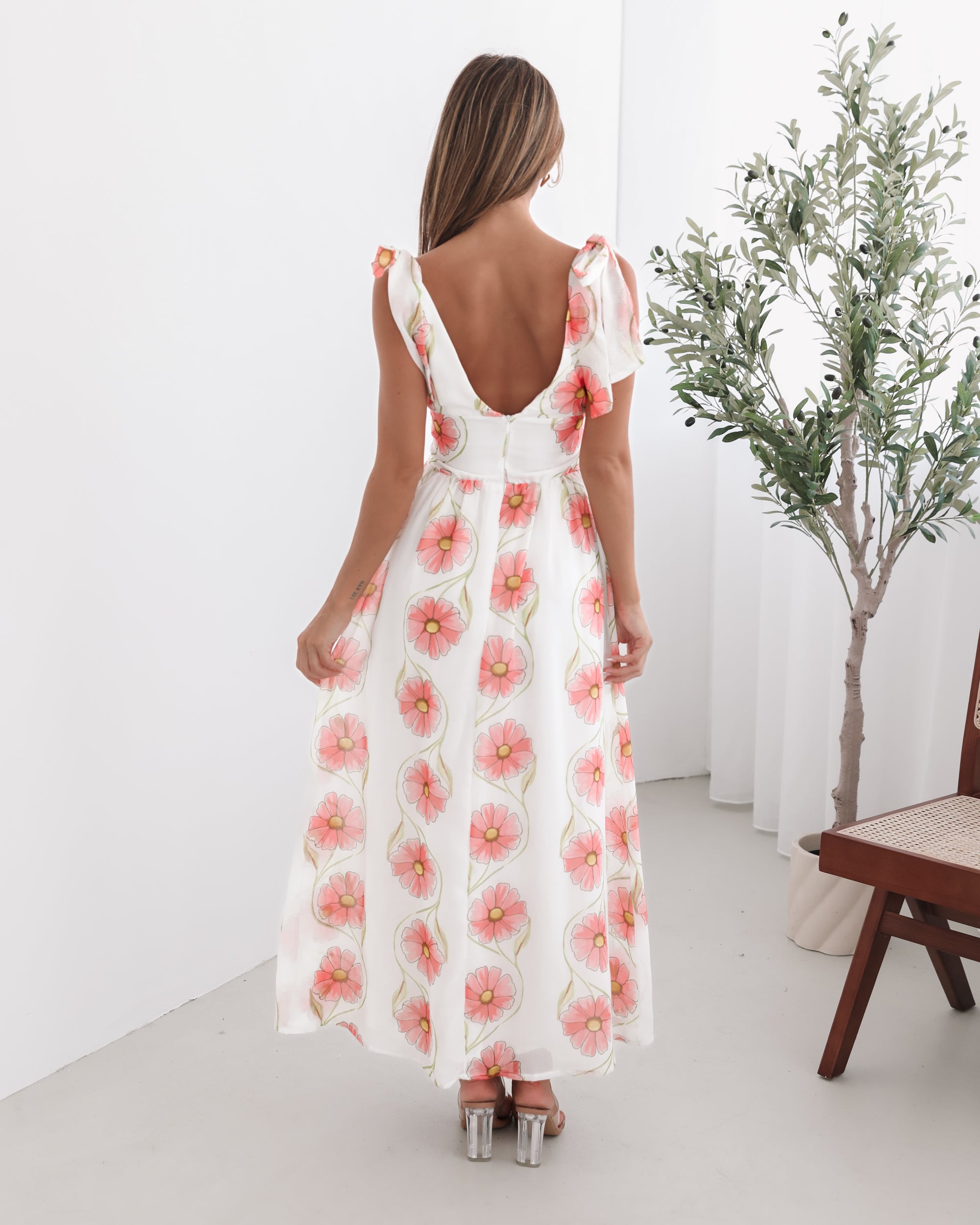 A woman standing in a room with a white floral maxi dress with bow straps and a plunge neckline.