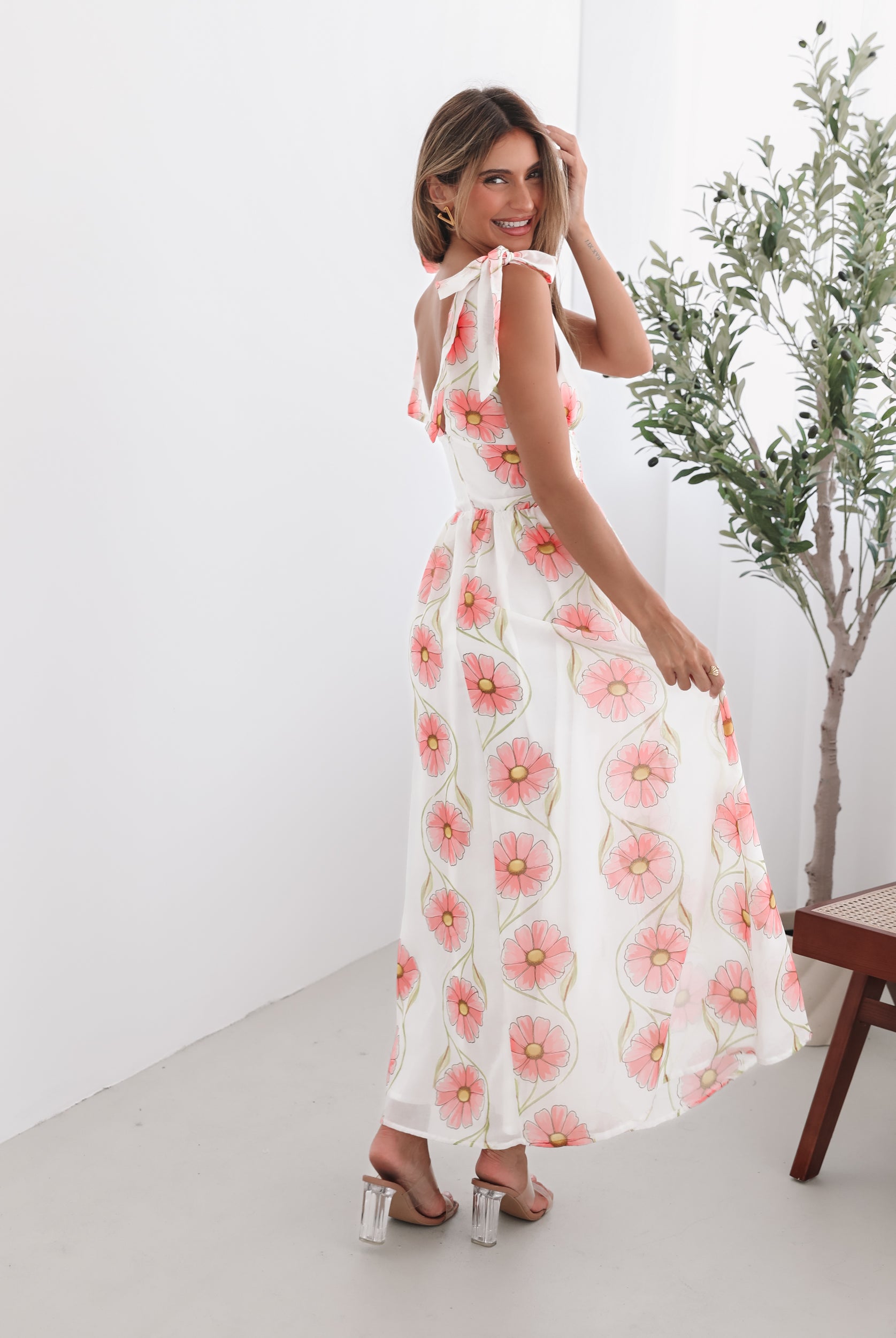A woman standing in a room with a white floral maxi dress with bow straps and a plunge neckline.