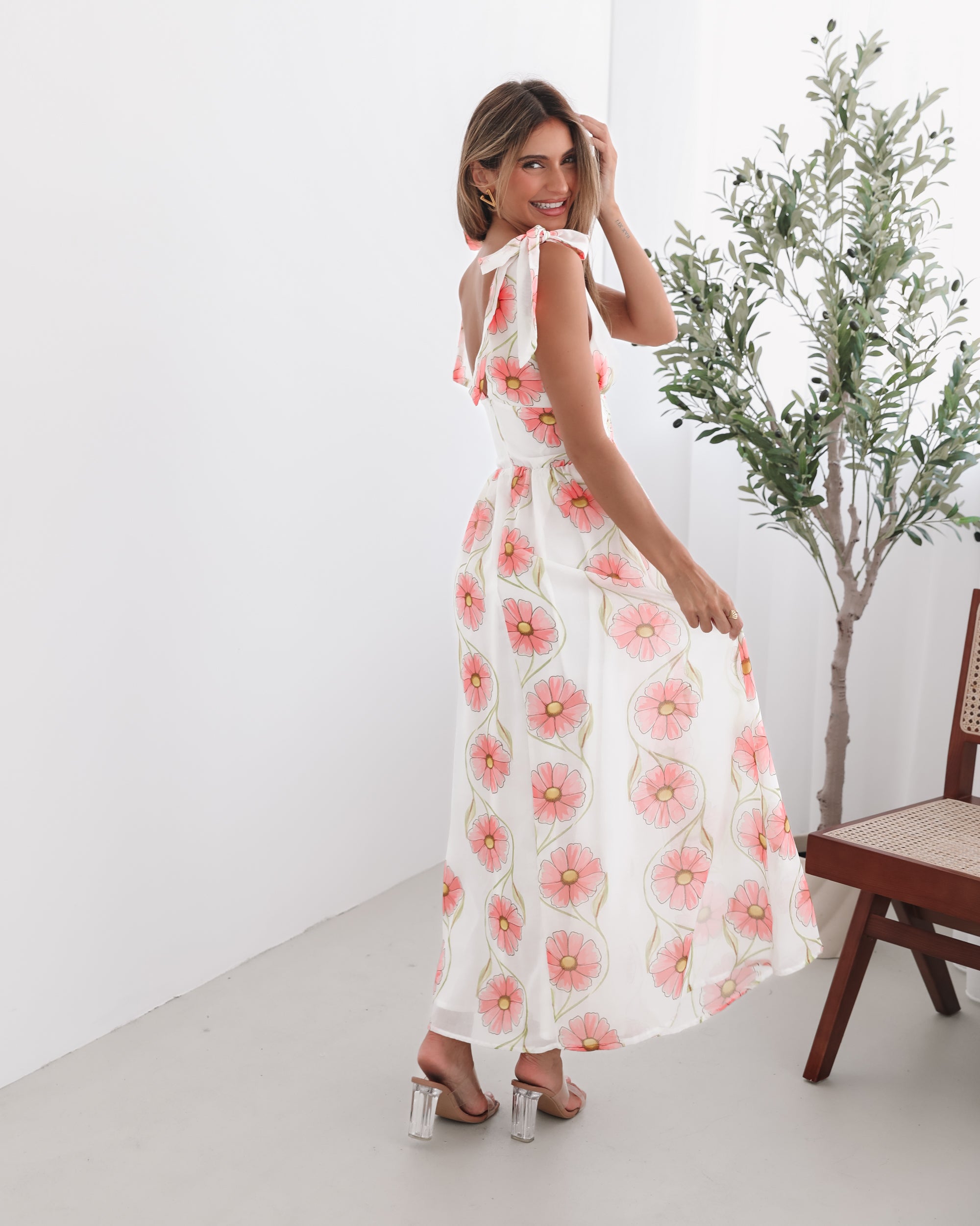A woman standing in a room with a white floral maxi dress with bow straps and a plunge neckline.