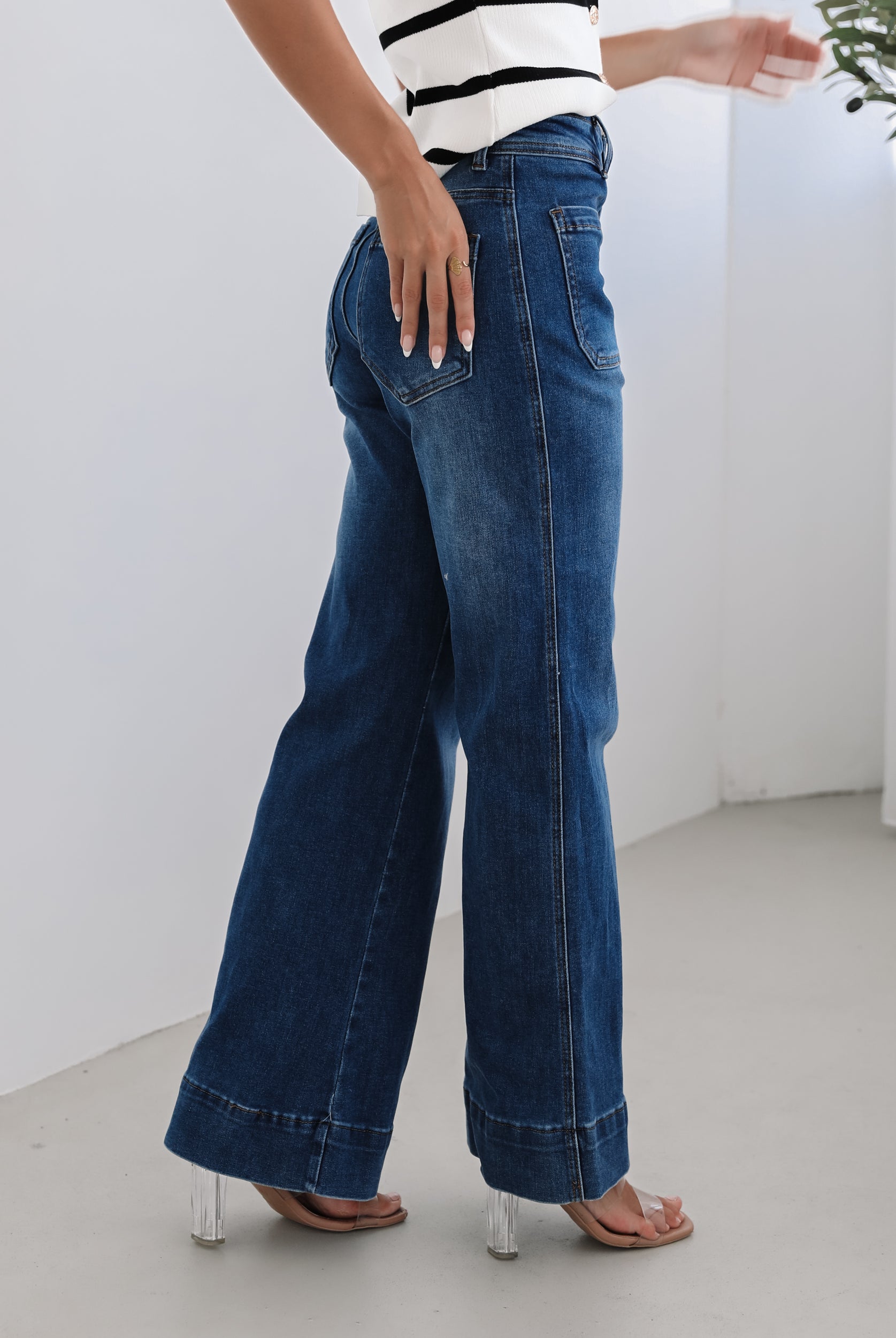 A woman standing in a room with light decor, wearing a striped top and mid-blue Yzabel Jeans with a high waist fit and wide leg silhouette.