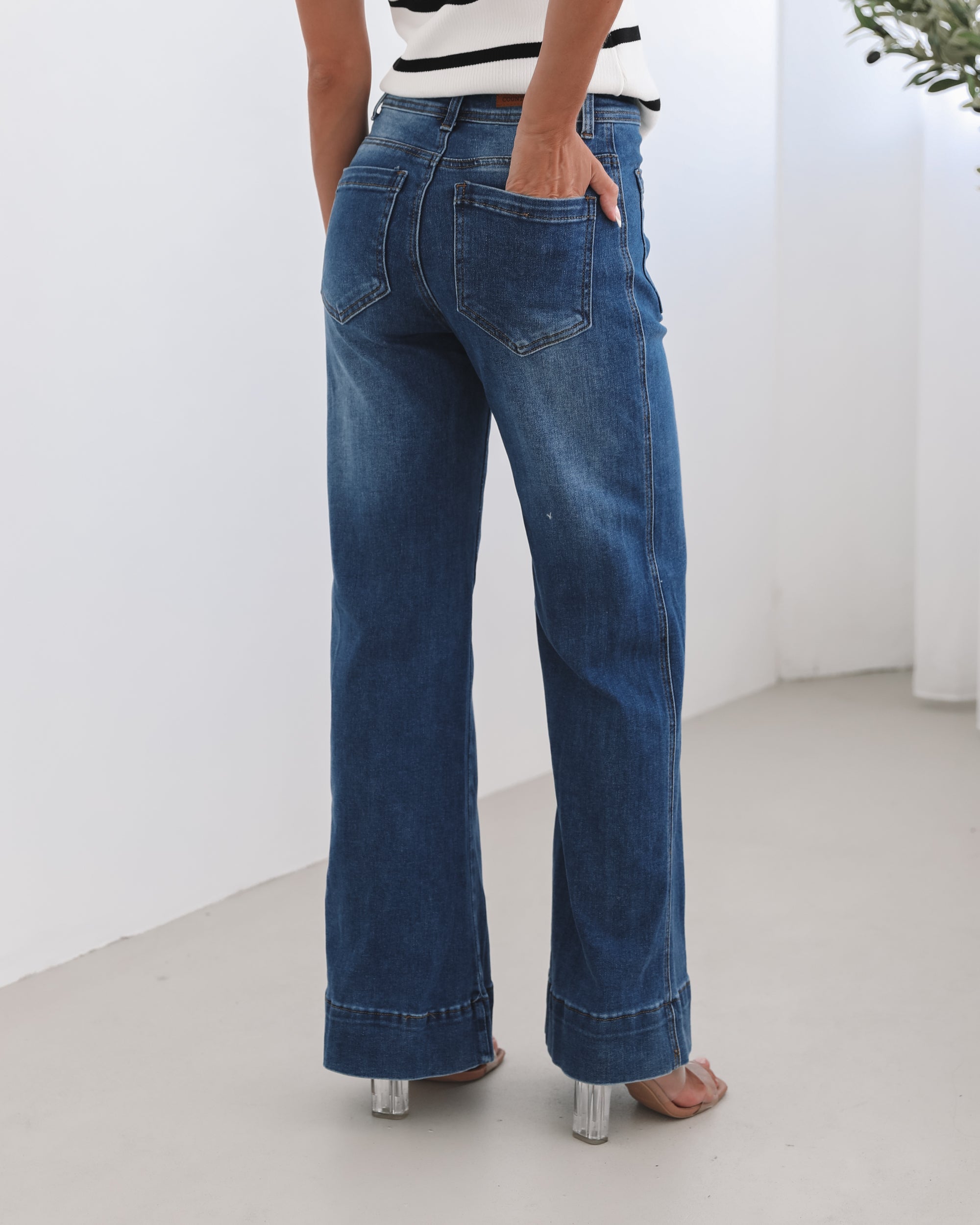 A woman standing in a room with light decor, wearing a striped top and mid-blue Yzabel Jeans with a high waist fit and wide leg silhouette.