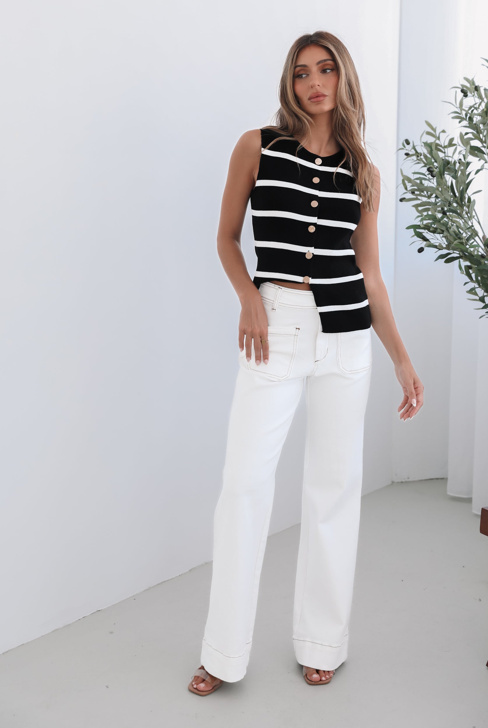 A female model wearing white high-waisted jeans with brown contrasting stitches, paired with a striped black and white top.