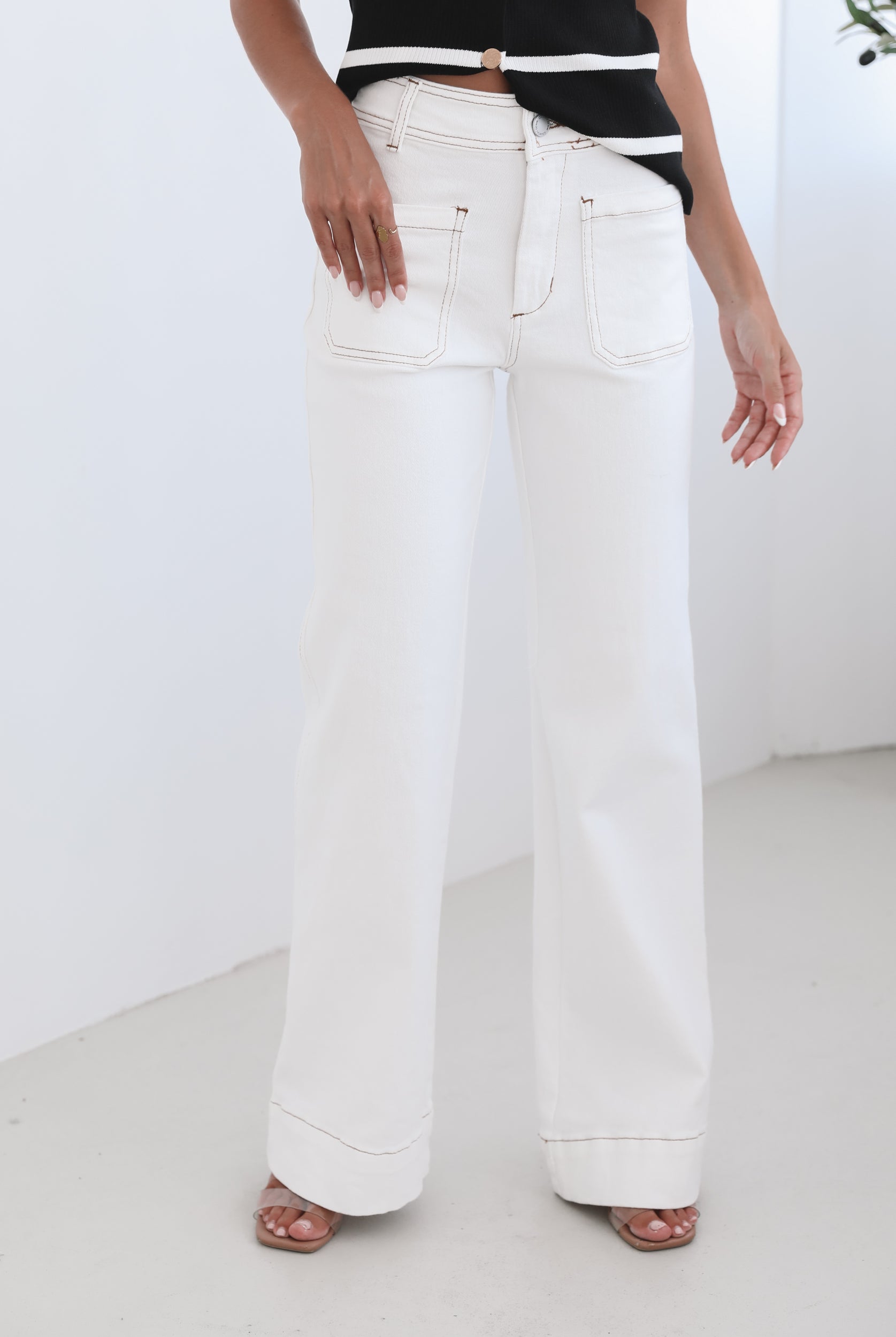 A female model wearing white high-waisted jeans with brown contrasting stitches, paired with a striped black and white top.