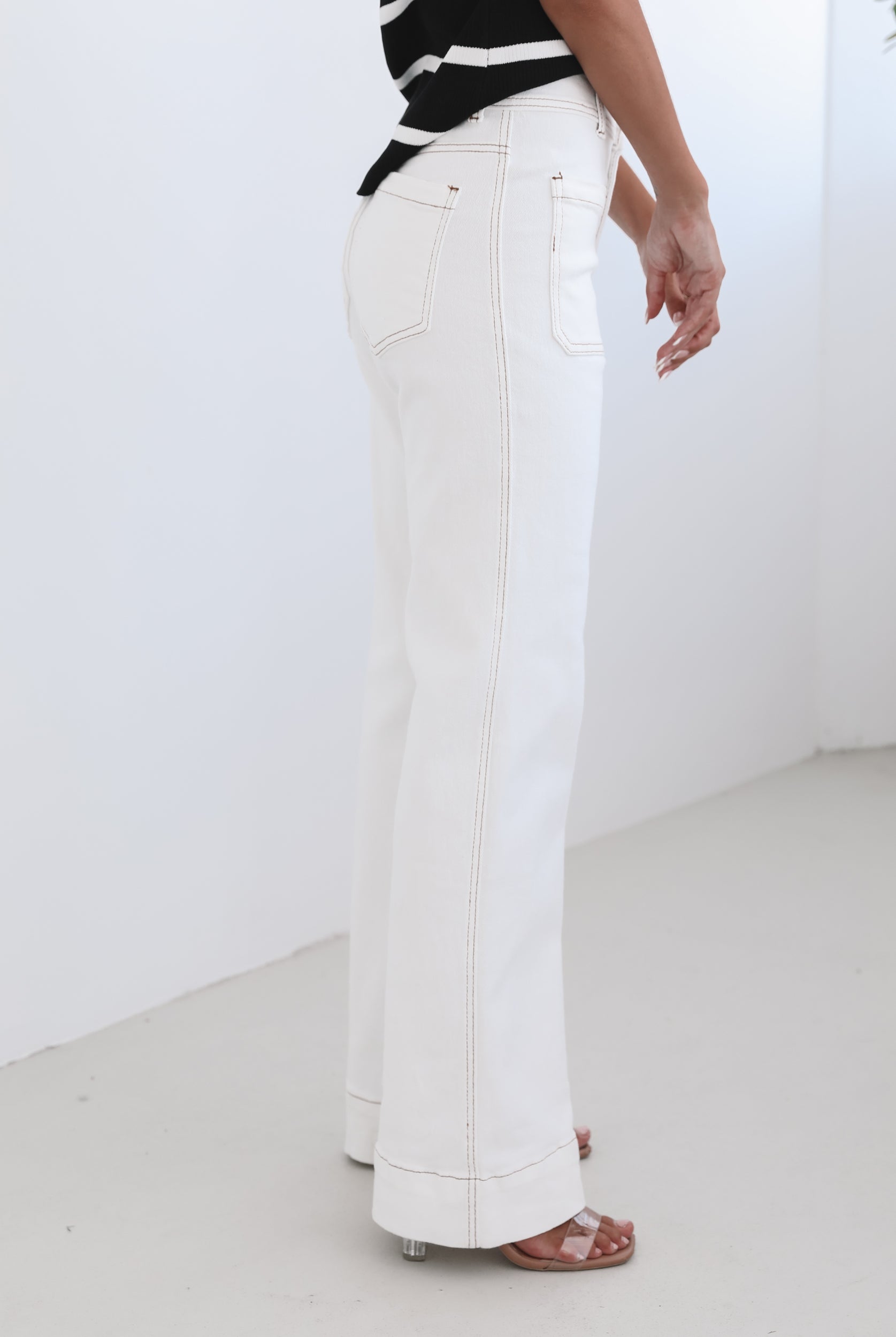 A female model wearing white high-waisted jeans with brown contrasting stitches, paired with a striped black and white top.