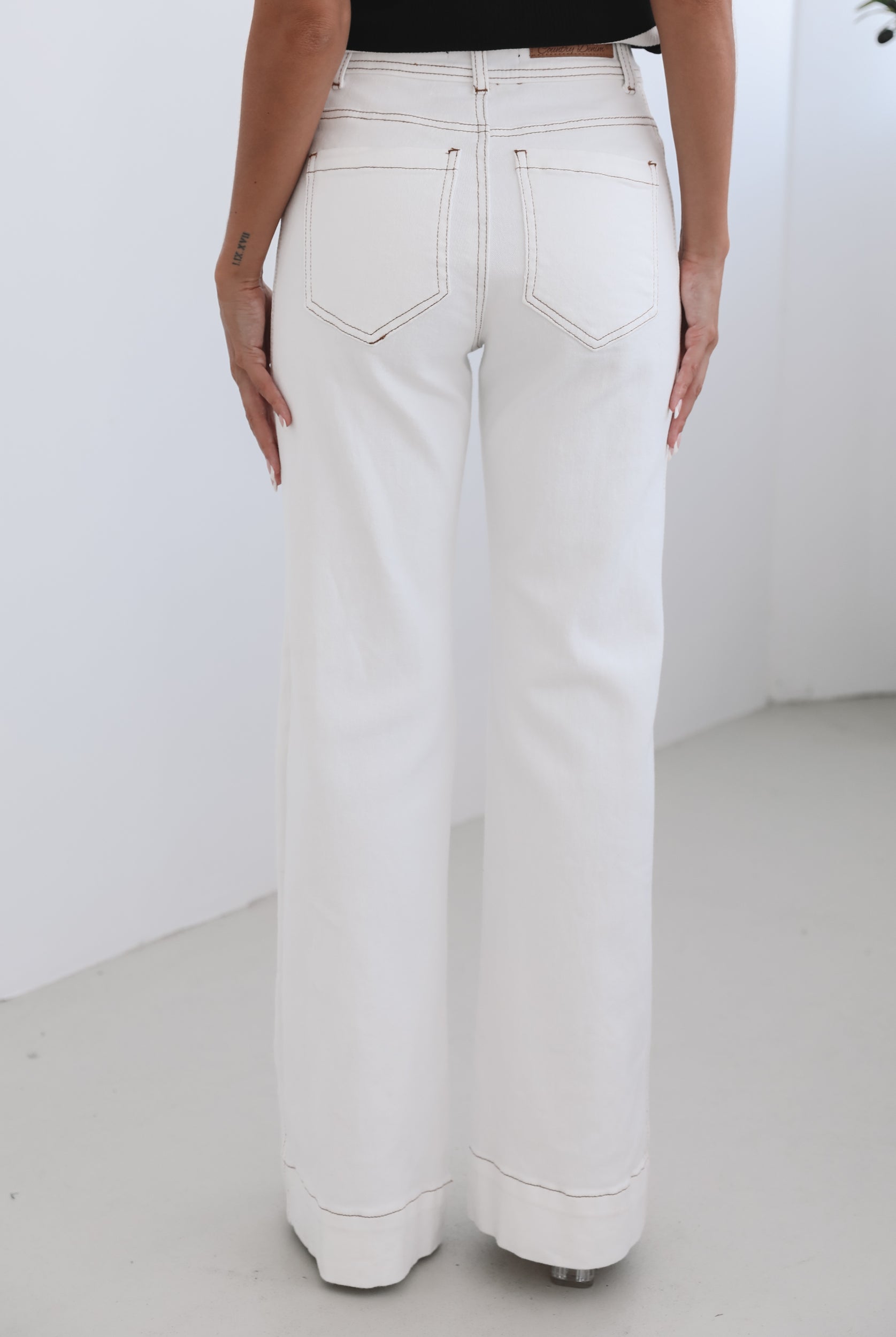 A female model wearing white high-waisted jeans with brown contrasting stitches, paired with a striped black and white top.