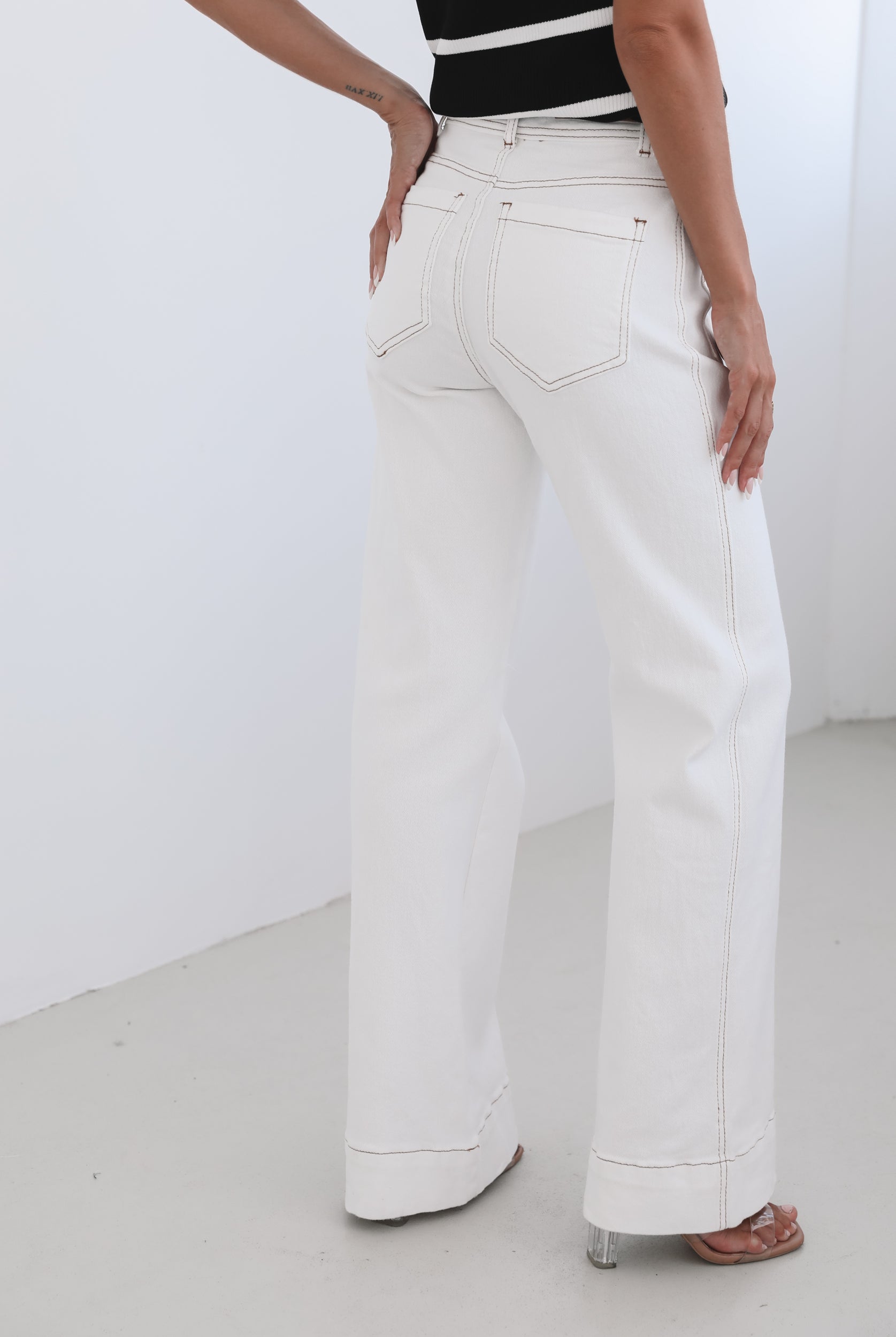 A female model wearing white high-waisted jeans with brown contrasting stitches, paired with a striped black and white top.