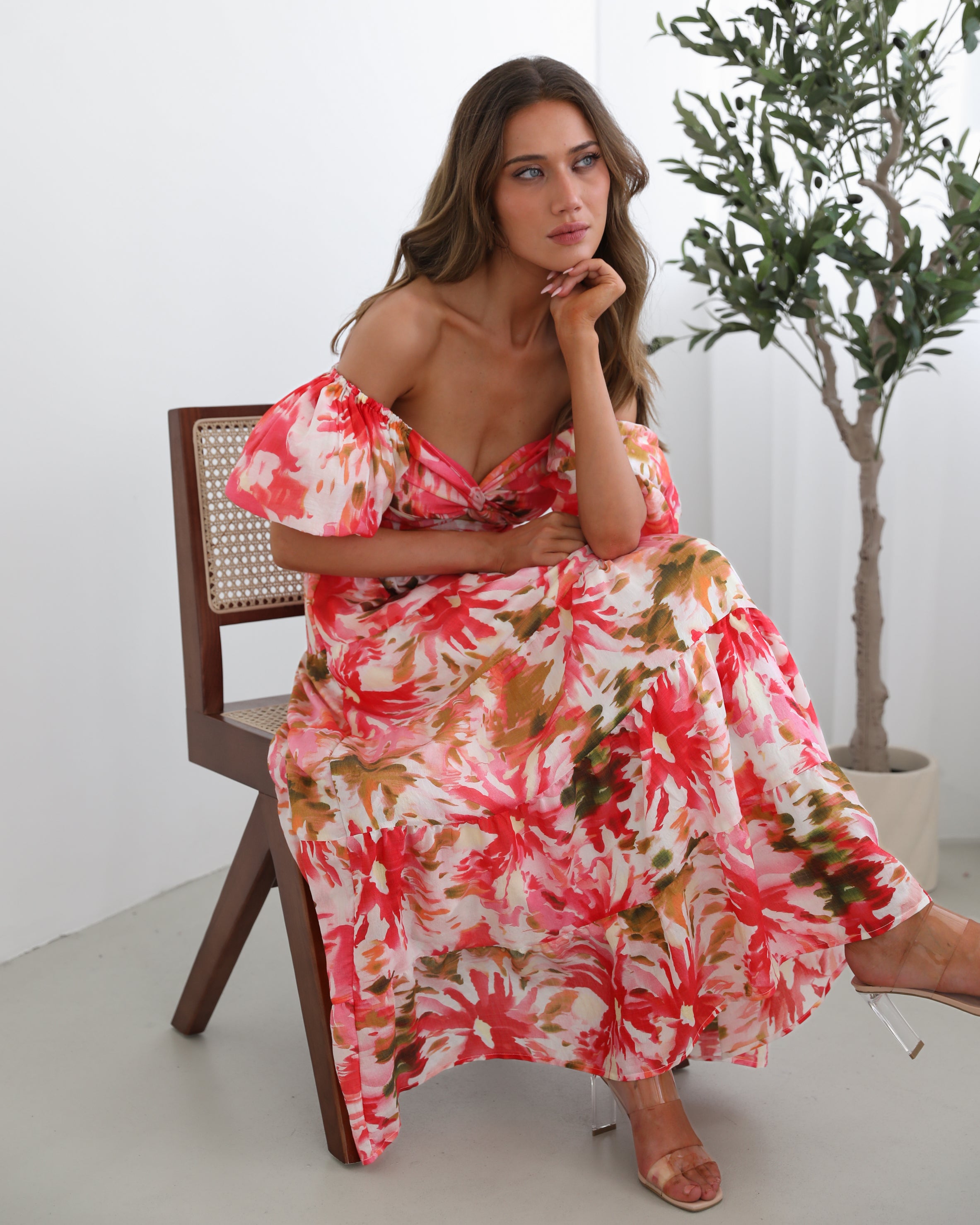A woman standing next to a chair, wearing a floral print maxi dress with a sweetheart neckline and short balloon sleeves. She is posing in a room with a large window and a plant in the background.