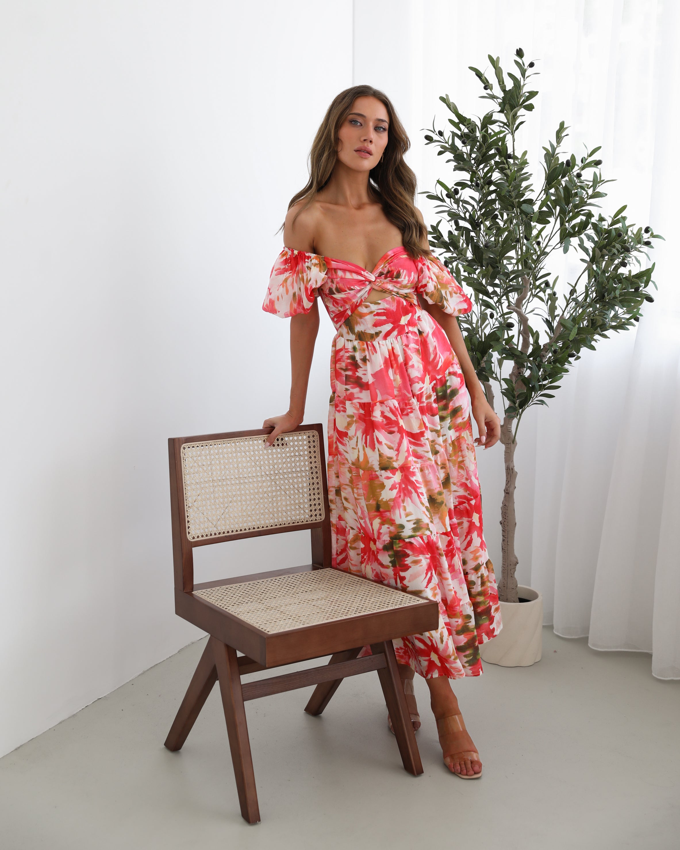 A woman standing next to a chair, wearing a floral print maxi dress with a sweetheart neckline and short balloon sleeves. She is posing in a room with a large window and a plant in the background.