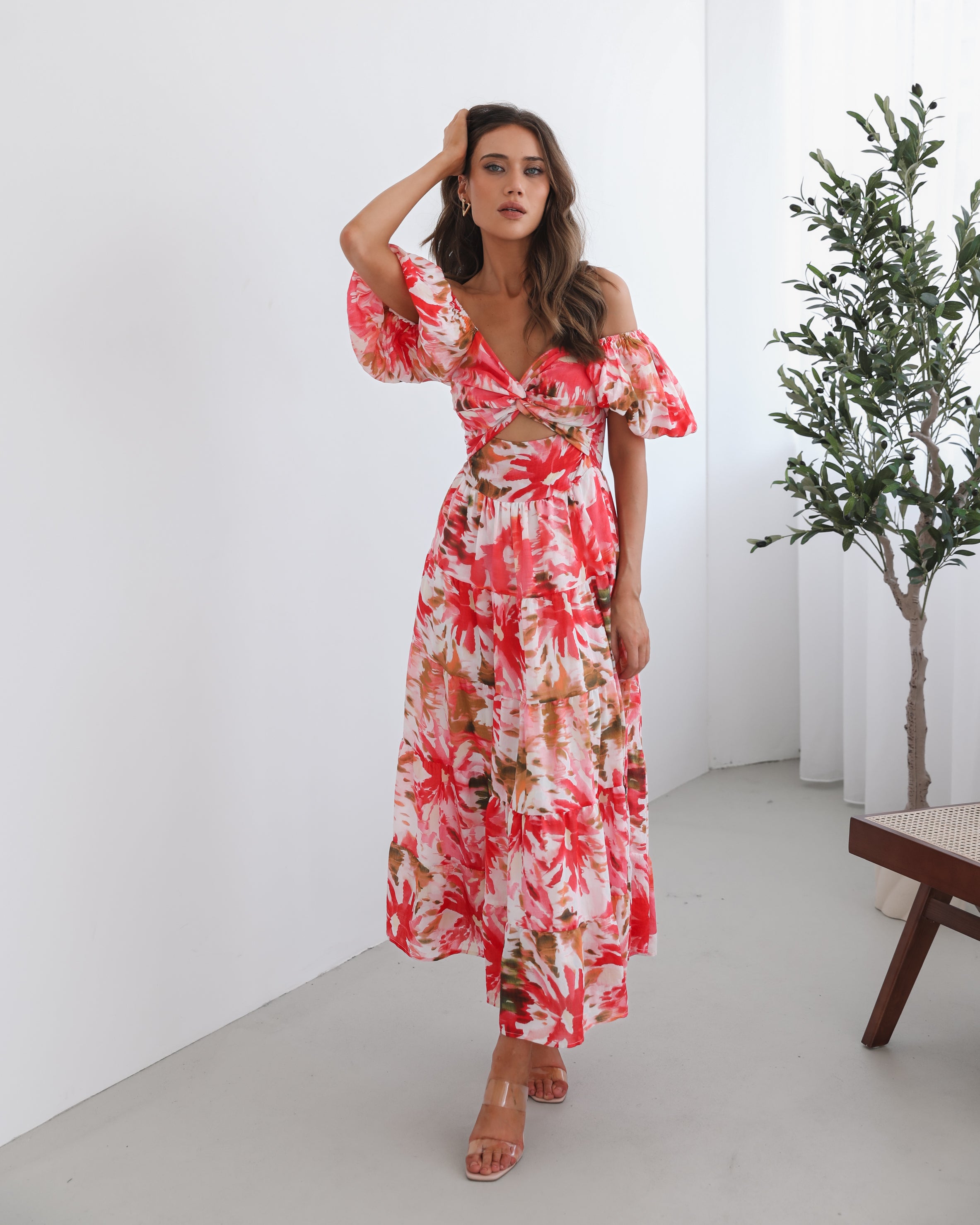 A woman standing next to a chair, wearing a floral print maxi dress with a sweetheart neckline and short balloon sleeves. She is posing in a room with a large window and a plant in the background.