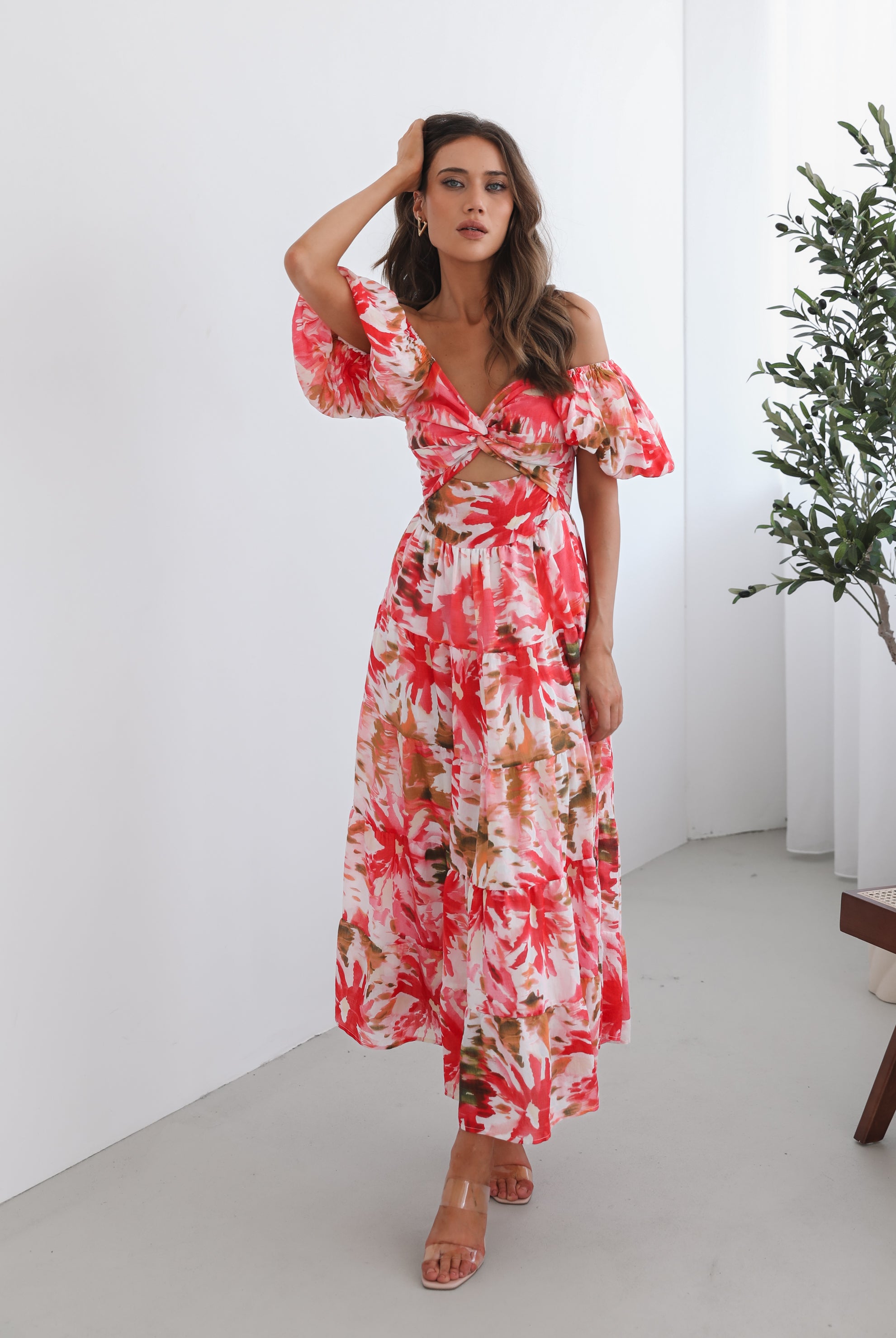 A woman standing next to a chair, wearing a floral print maxi dress with a sweetheart neckline and short balloon sleeves. She is posing in a room with a large window and a plant in the background.