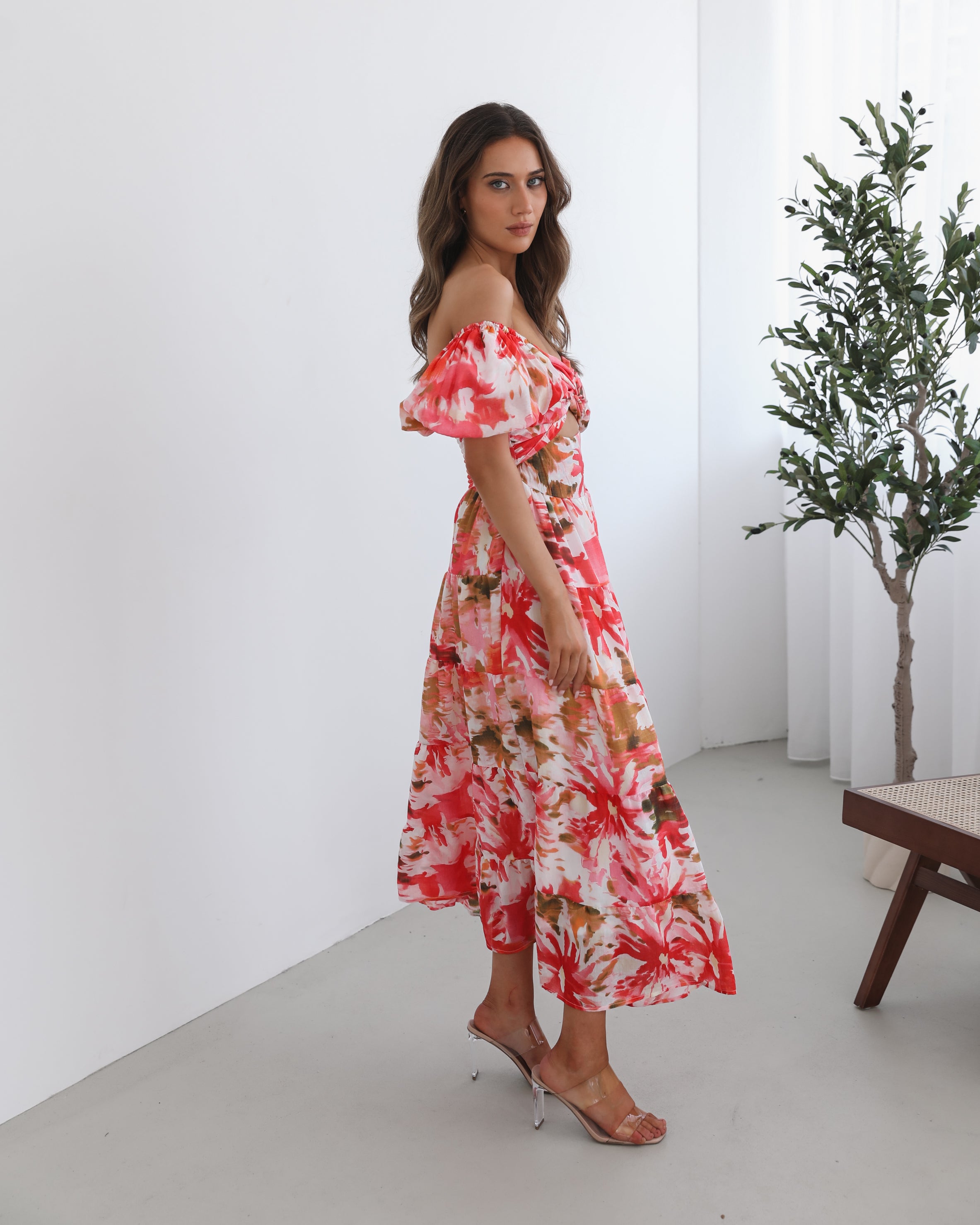 A woman standing next to a chair, wearing a floral print maxi dress with a sweetheart neckline and short balloon sleeves. She is posing in a room with a large window and a plant in the background.