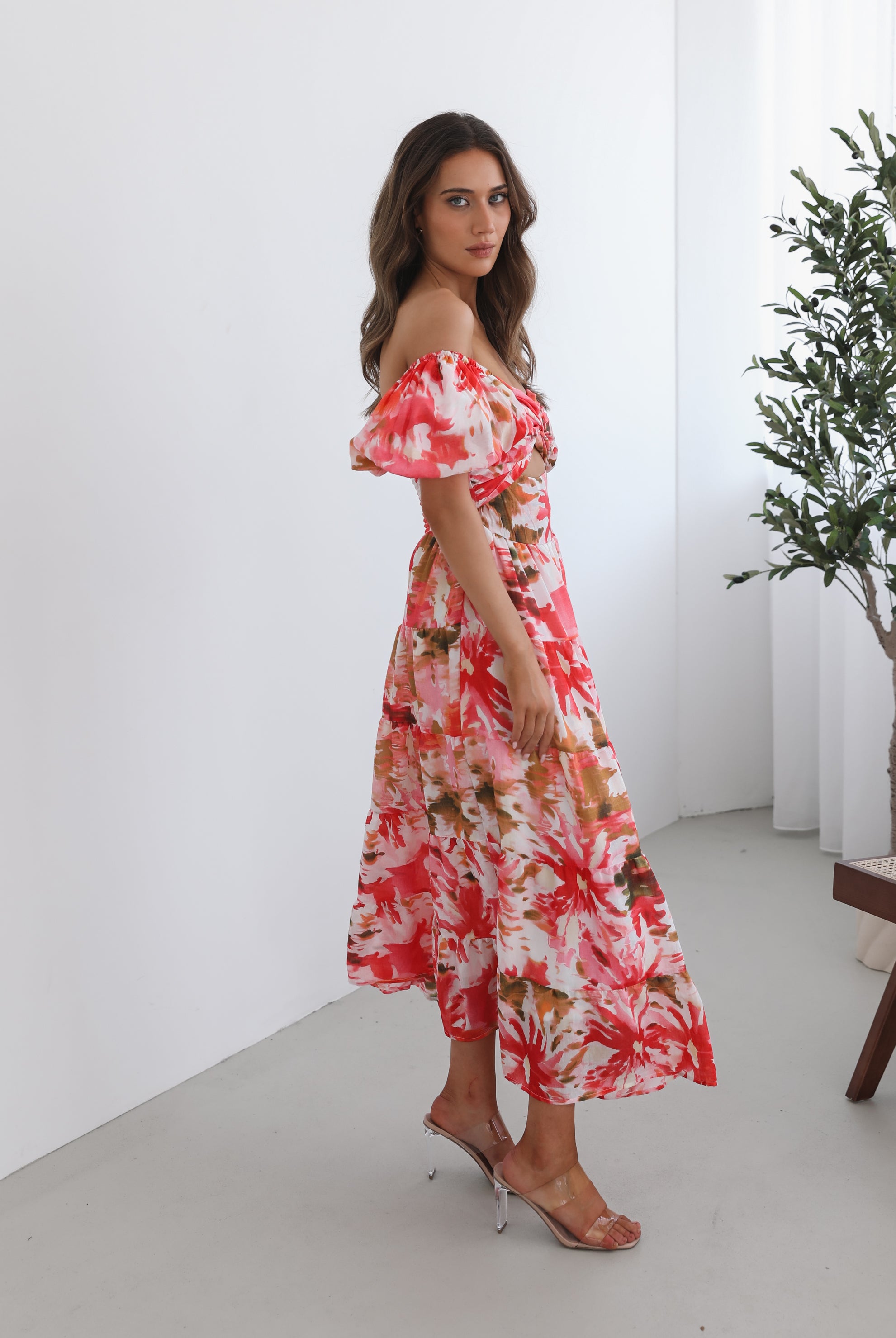 A woman standing next to a chair, wearing a floral print maxi dress with a sweetheart neckline and short balloon sleeves. She is posing in a room with a large window and a plant in the background.