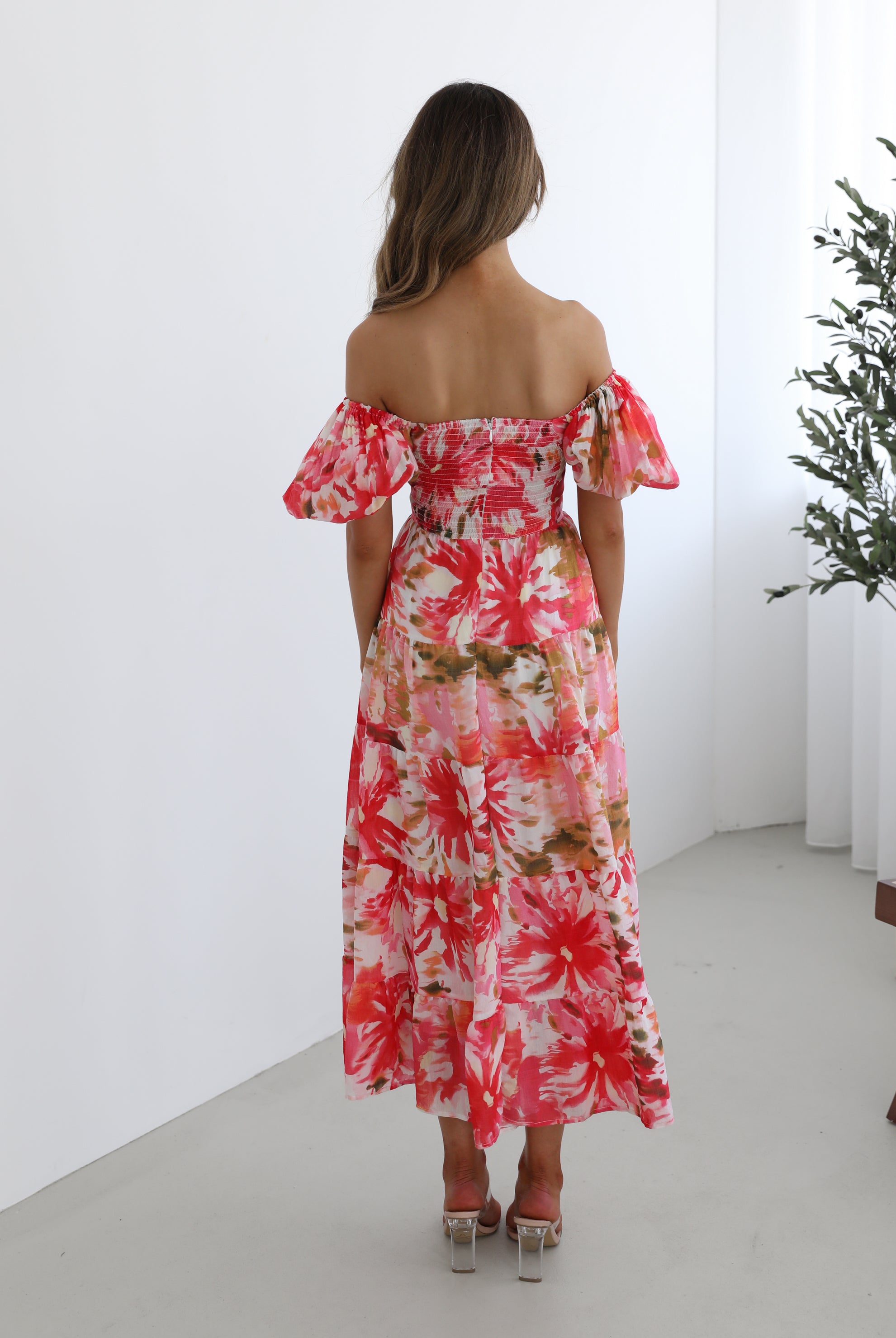 A woman standing next to a chair, wearing a floral print maxi dress with a sweetheart neckline and short balloon sleeves. She is posing in a room with a large window and a plant in the background.