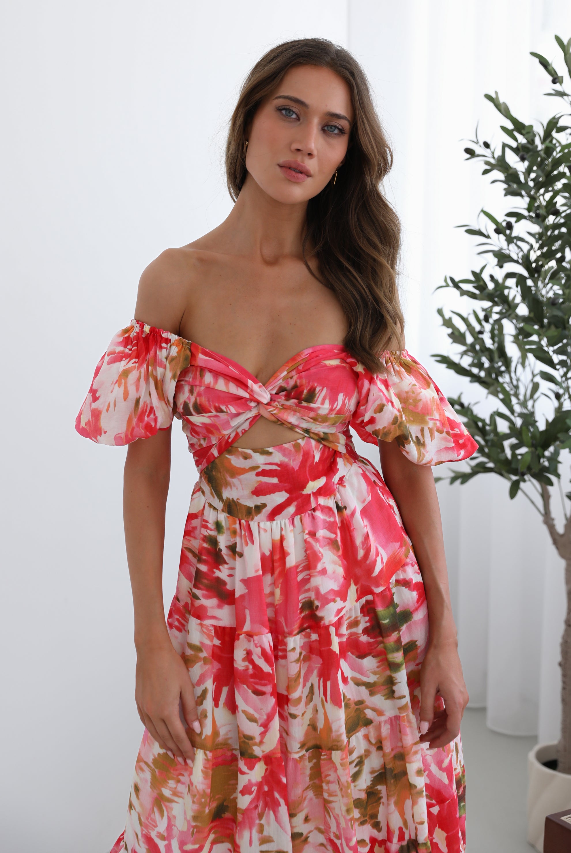 A woman standing next to a chair, wearing a floral print maxi dress with a sweetheart neckline and short balloon sleeves. She is posing in a room with a large window and a plant in the background.