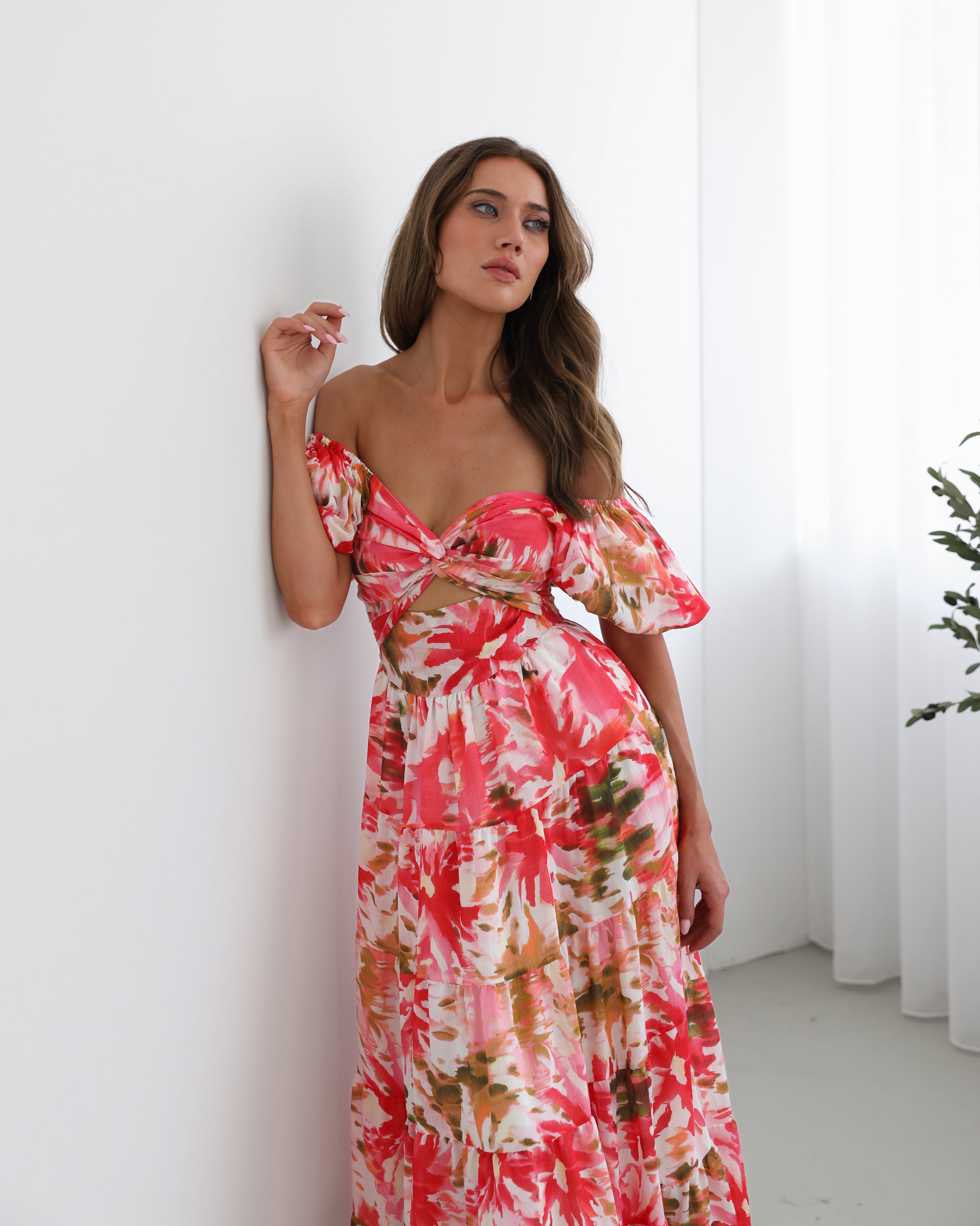 A woman standing next to a chair, wearing a floral print maxi dress with a sweetheart neckline and short balloon sleeves. She is posing in a room with a large window and a plant in the background.