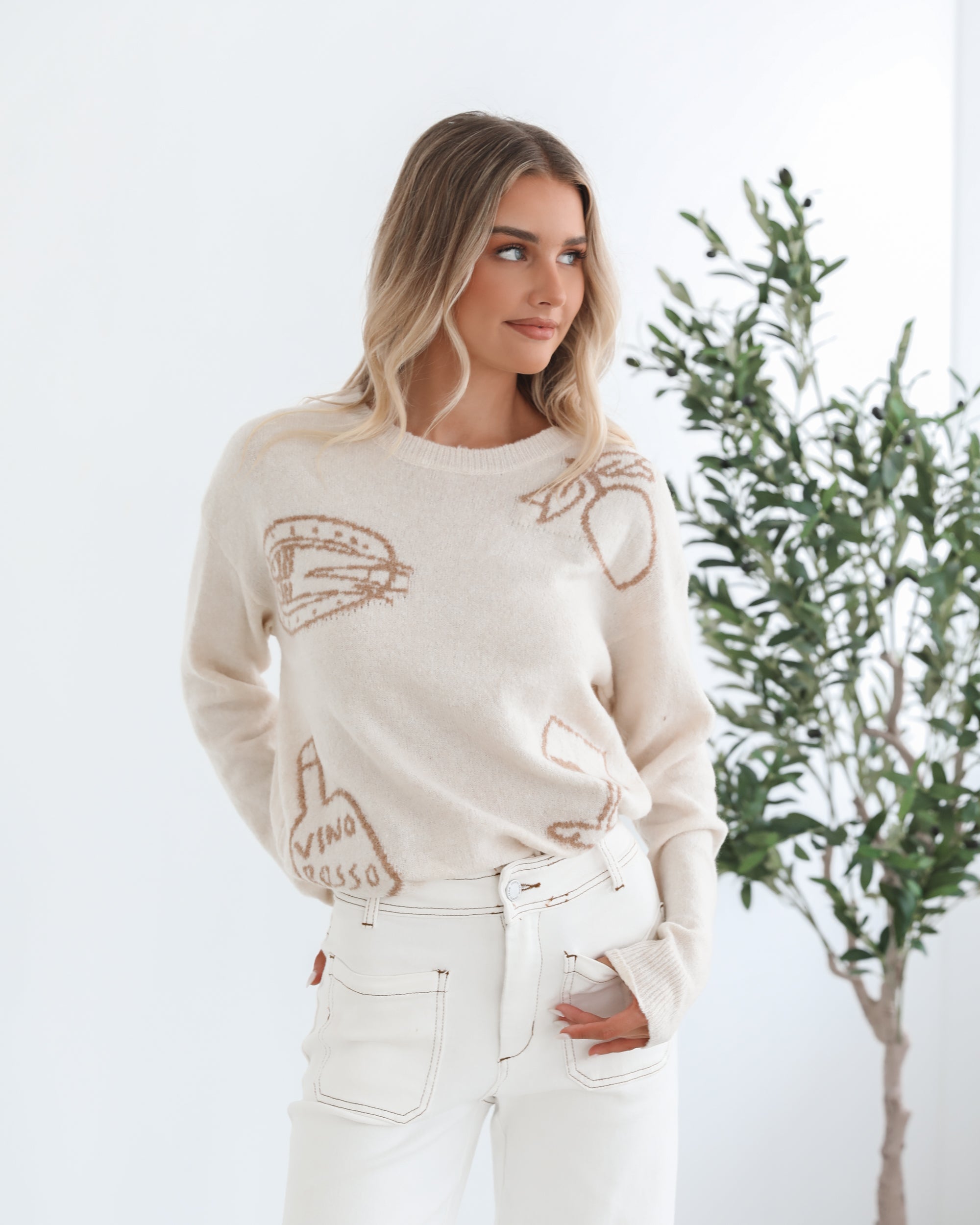 A woman wearing a cream-colored knit top with a round neckline and long sleeves, featuring a print of various items.