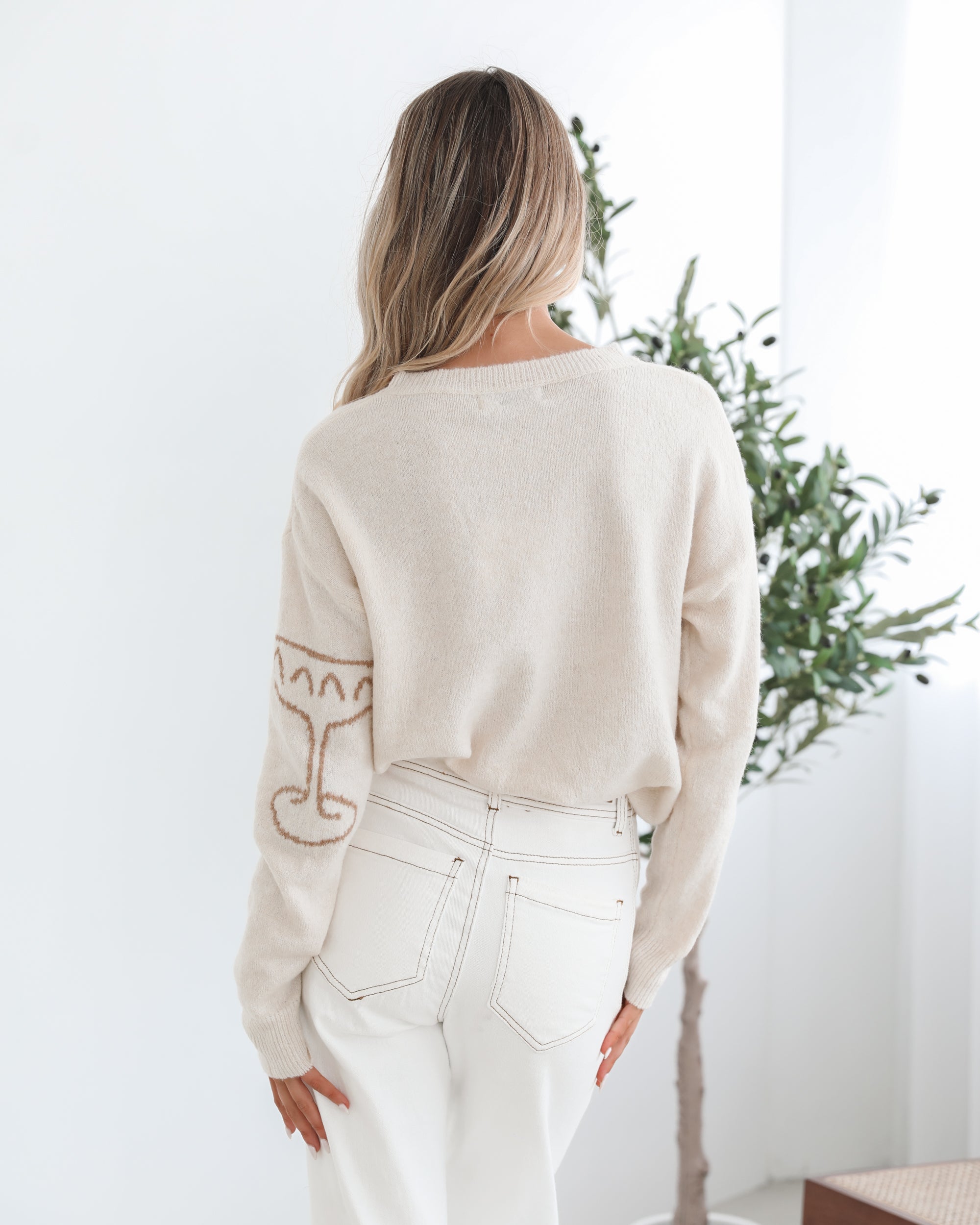 A woman wearing a cream-colored knit top with a round neckline and long sleeves, featuring a print of various items.