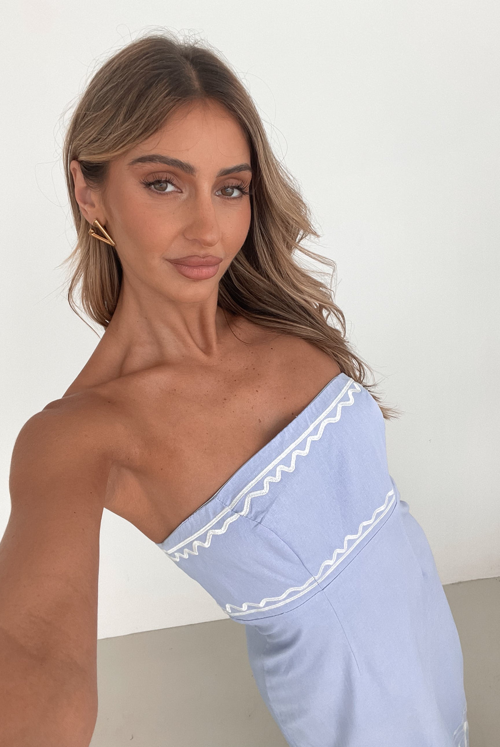 A woman wearing a blue and white floral print maxi dress with a tube style top and fit-to-flare silhouette. She is standing in a room with a white wall and a wooden bench, posing with one hand on her hip.