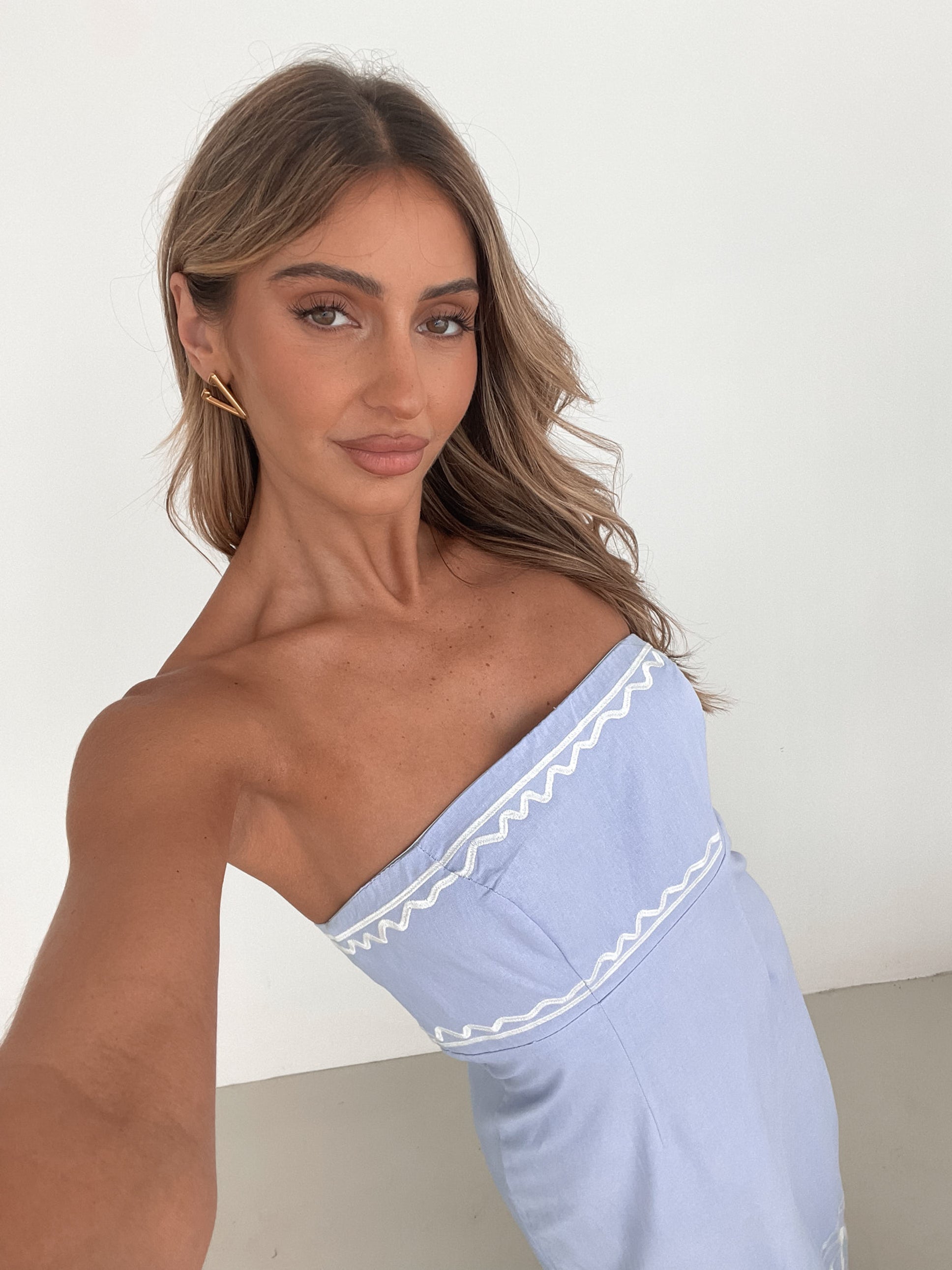 A woman wearing a blue and white floral print maxi dress with a tube style top and fit-to-flare silhouette. She is standing in a room with a white wall and a wooden bench, posing with one hand on her hip.