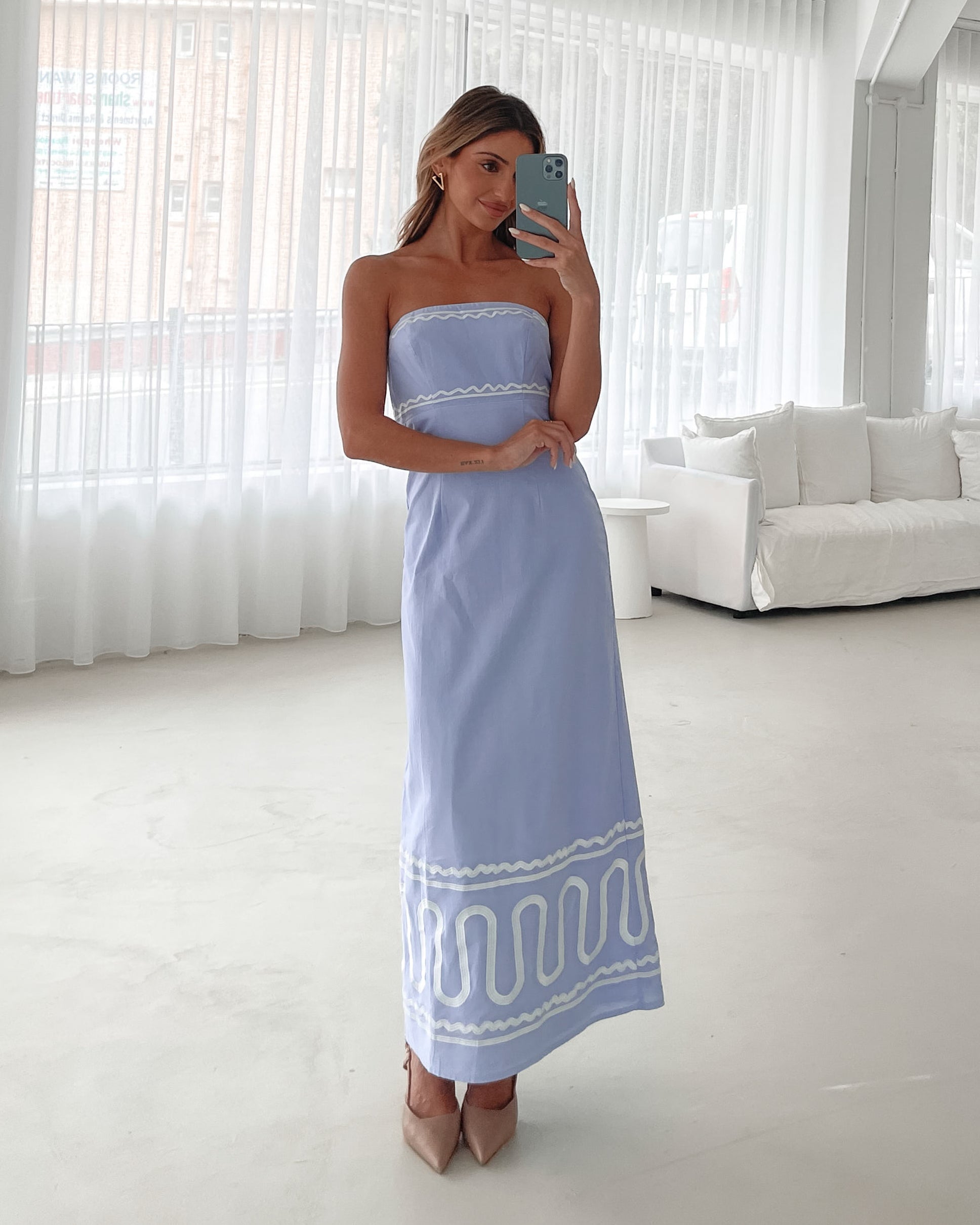 A woman wearing a blue and white floral print maxi dress with a tube style top and fit-to-flare silhouette. She is standing in a room with a white wall and a wooden bench, posing with one hand on her hip.