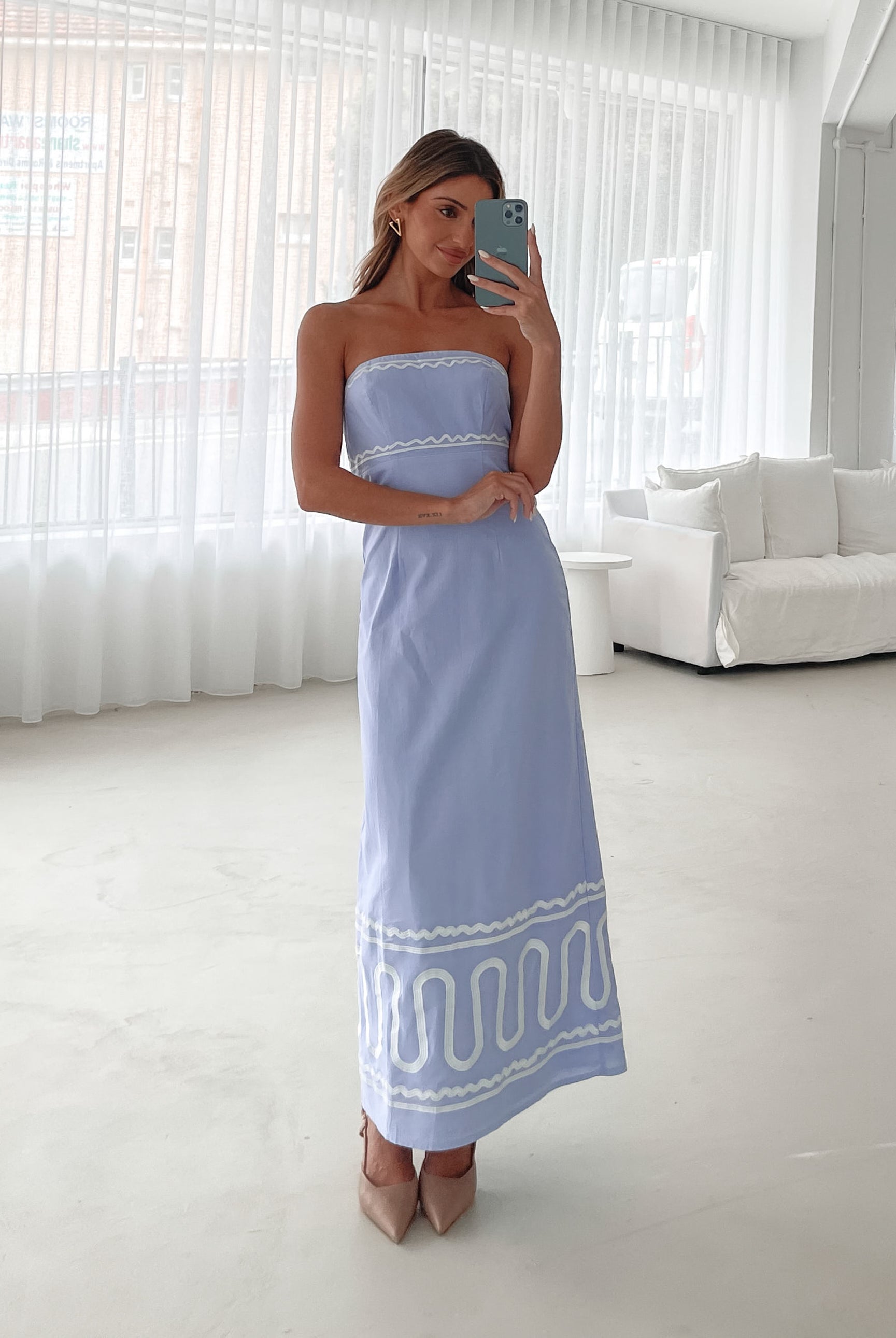 A woman wearing a blue and white floral print maxi dress with a tube style top and fit-to-flare silhouette. She is standing in a room with a white wall and a wooden bench, posing with one hand on her hip.