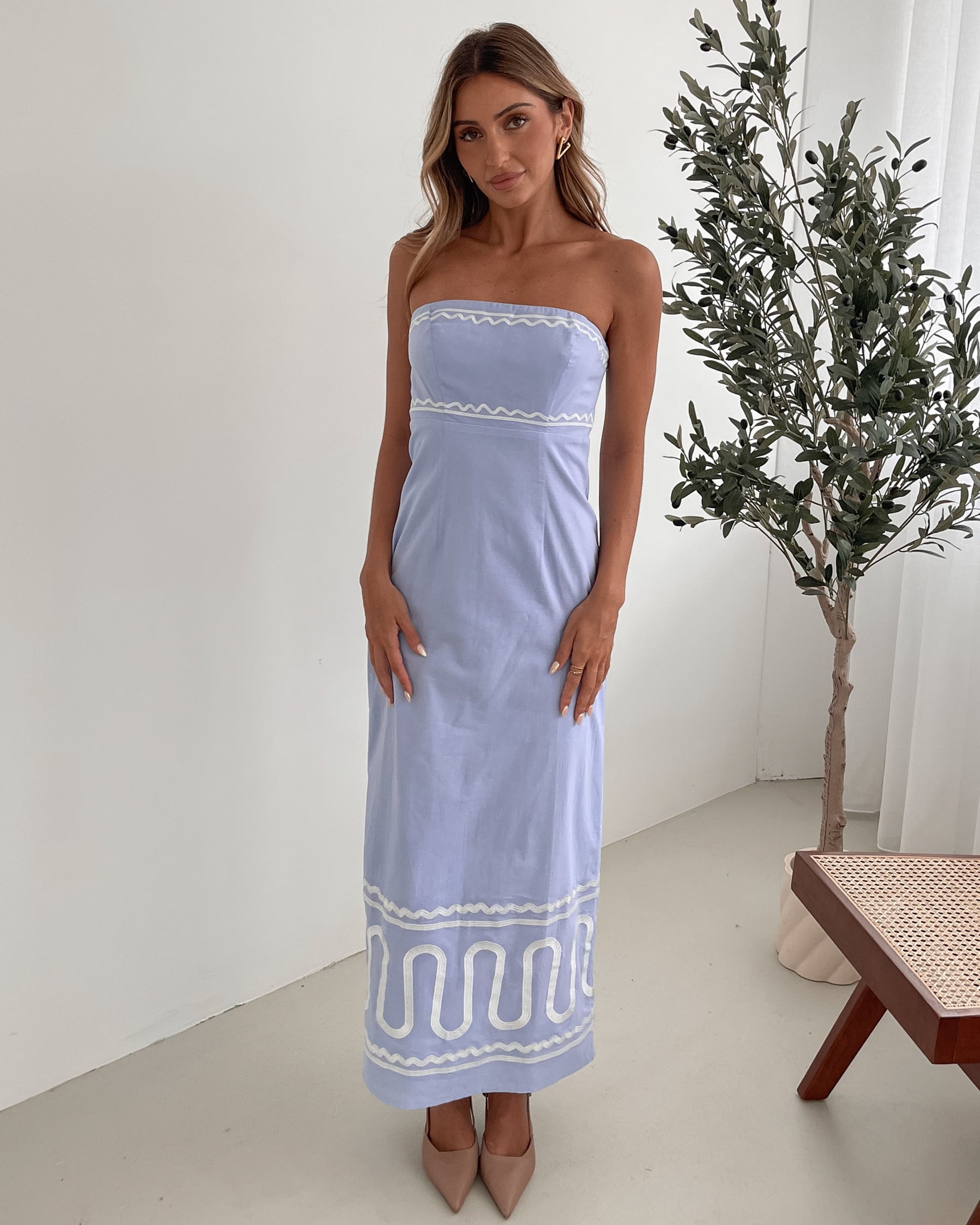 A woman wearing a blue and white floral print maxi dress with a tube style top and fit-to-flare silhouette. She is standing in a room with a white wall and a wooden bench, posing with one hand on her hip.