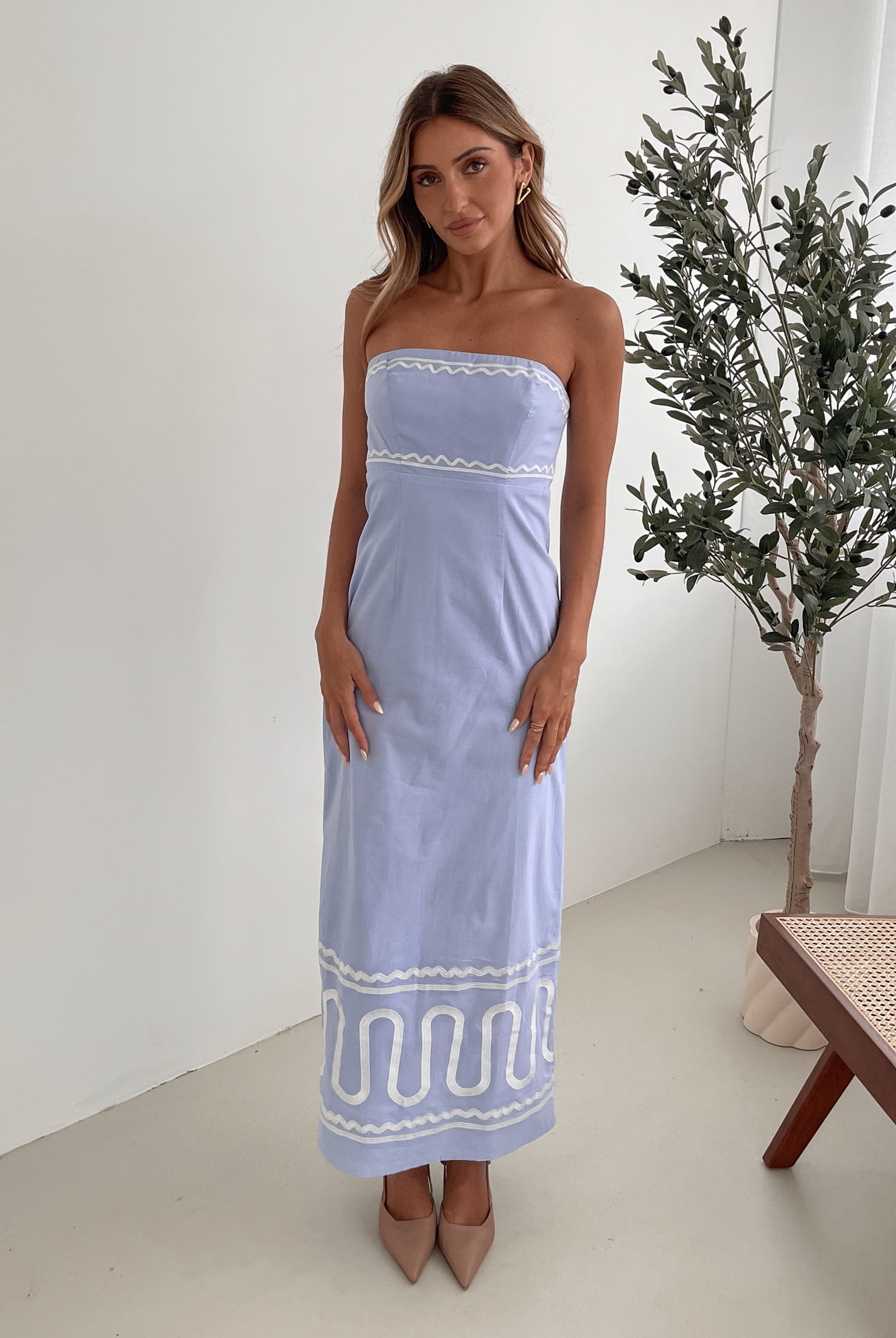 A woman wearing a blue and white floral print maxi dress with a tube style top and fit-to-flare silhouette. She is standing in a room with a white wall and a wooden bench, posing with one hand on her hip.
