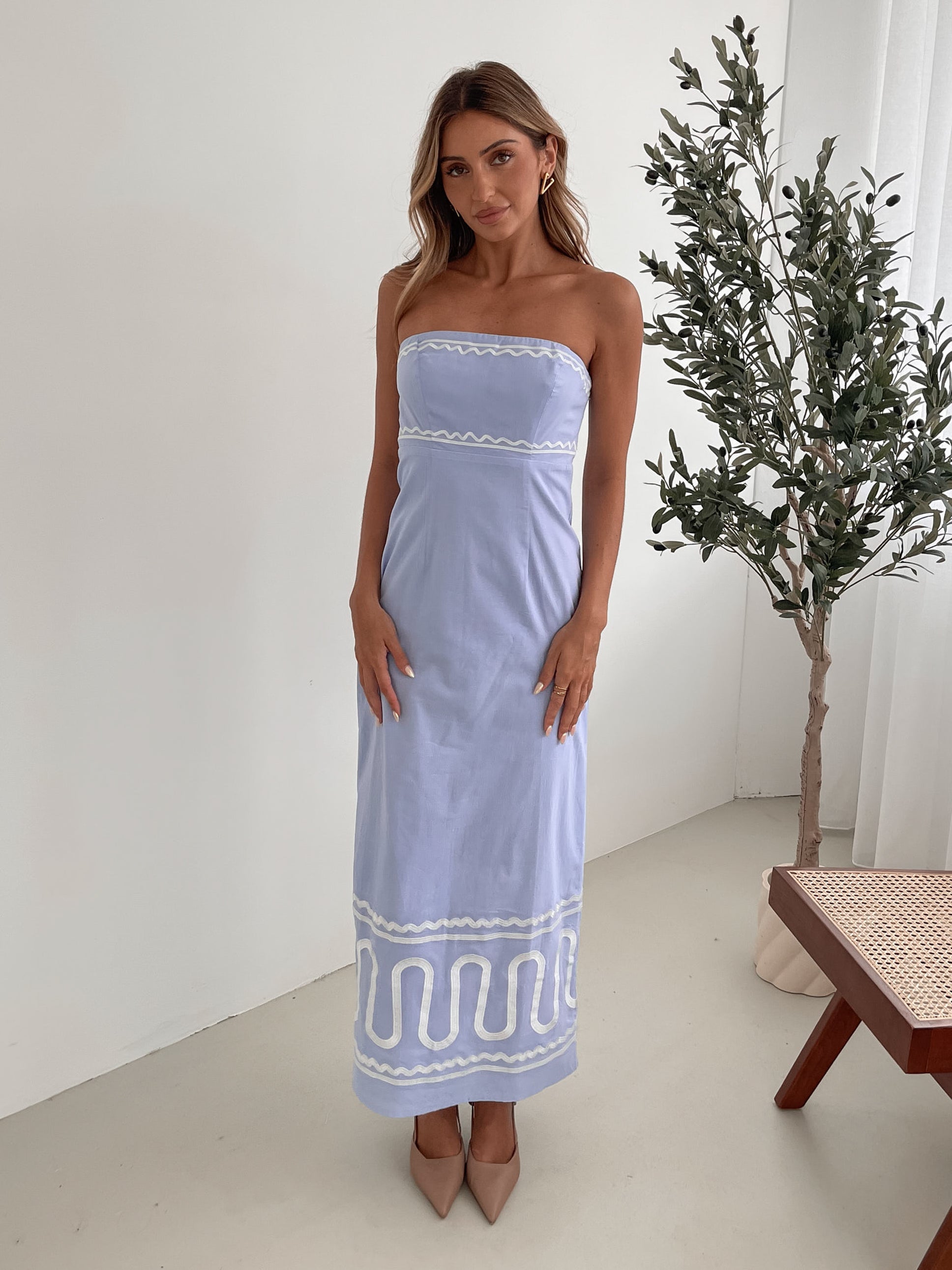 A woman wearing a blue and white floral print maxi dress with a tube style top and fit-to-flare silhouette. She is standing in a room with a white wall and a wooden bench, posing with one hand on her hip.