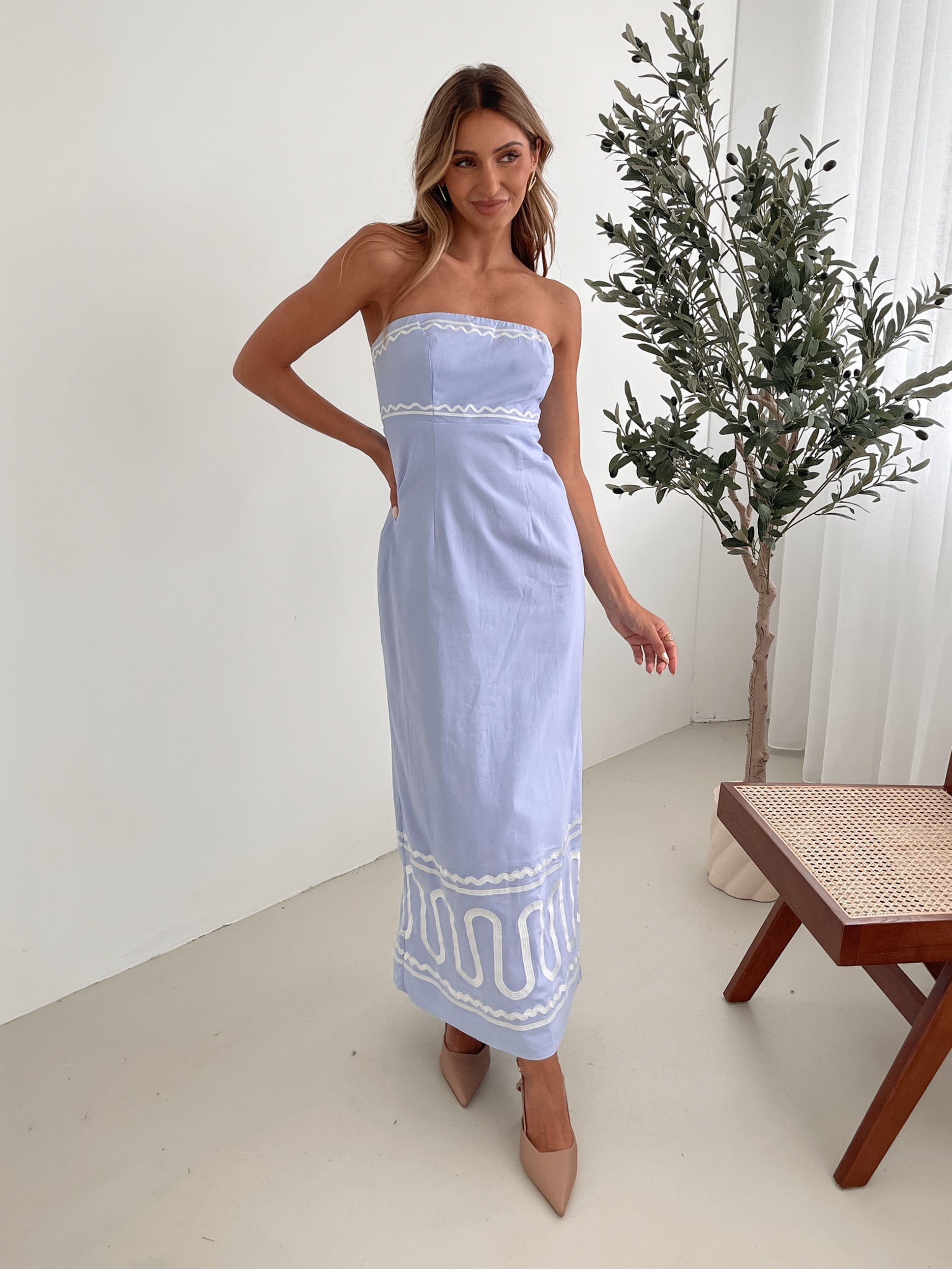 A woman wearing a blue and white floral print maxi dress with a tube style top and fit-to-flare silhouette. She is standing in a room with a white wall and a wooden bench, posing with one hand on her hip.