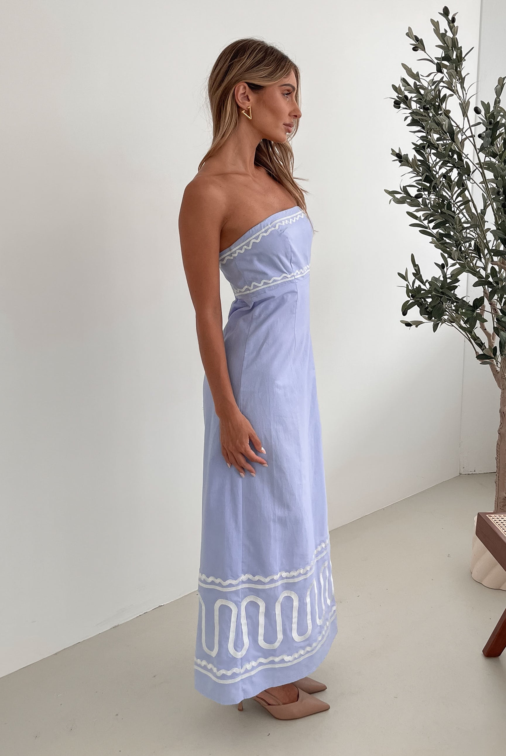 A woman wearing a blue and white floral print maxi dress with a tube style top and fit-to-flare silhouette. She is standing in a room with a white wall and a wooden bench, posing with one hand on her hip.