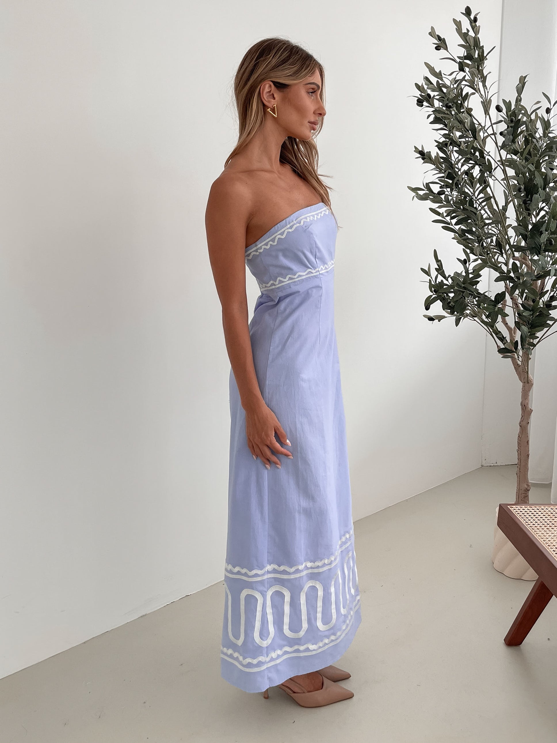 A woman wearing a blue and white floral print maxi dress with a tube style top and fit-to-flare silhouette. She is standing in a room with a white wall and a wooden bench, posing with one hand on her hip.