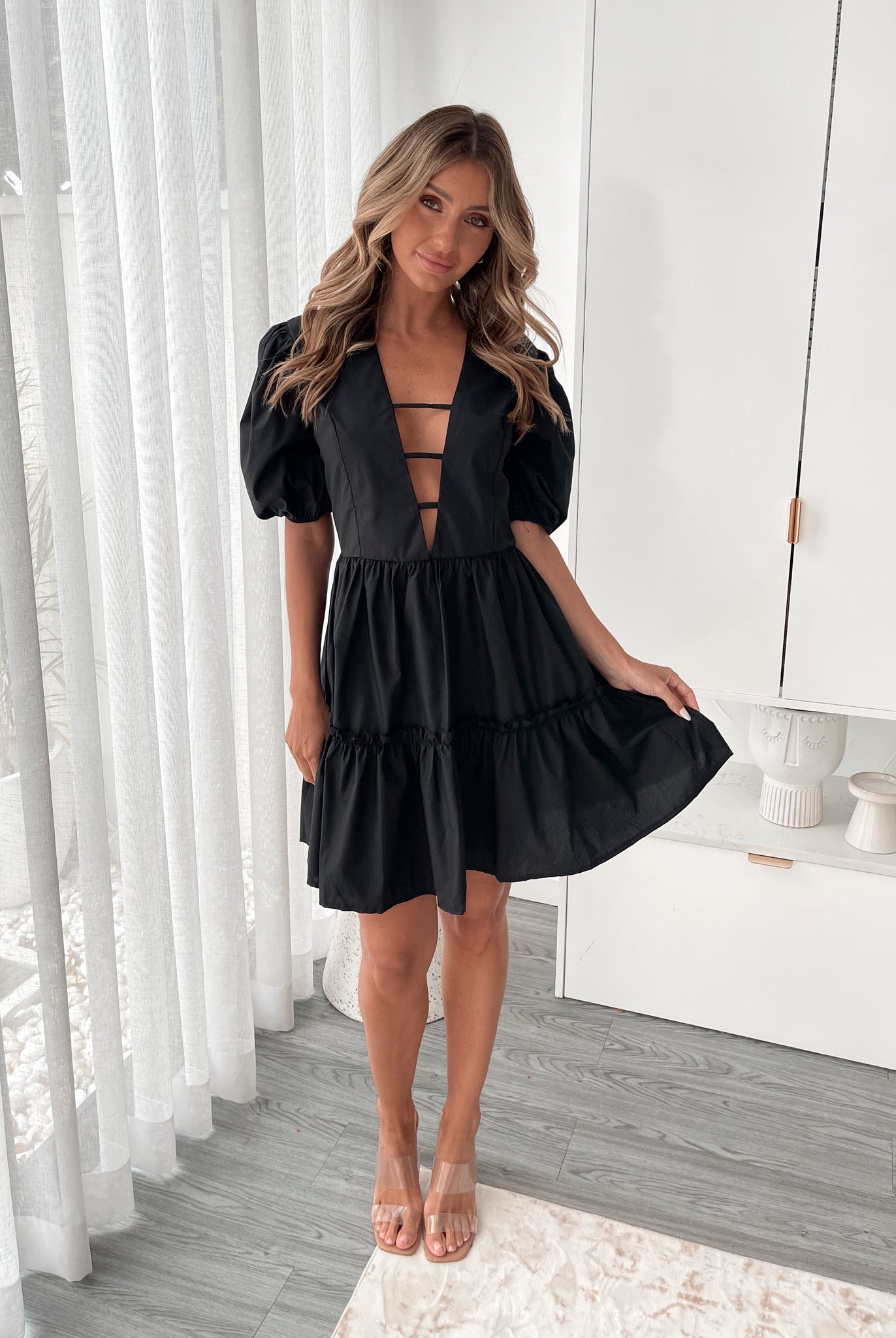 A woman wearing a classic black mini dress with a baby doll-inspired shape, featuring a plunge neckline and balloon sleeves.