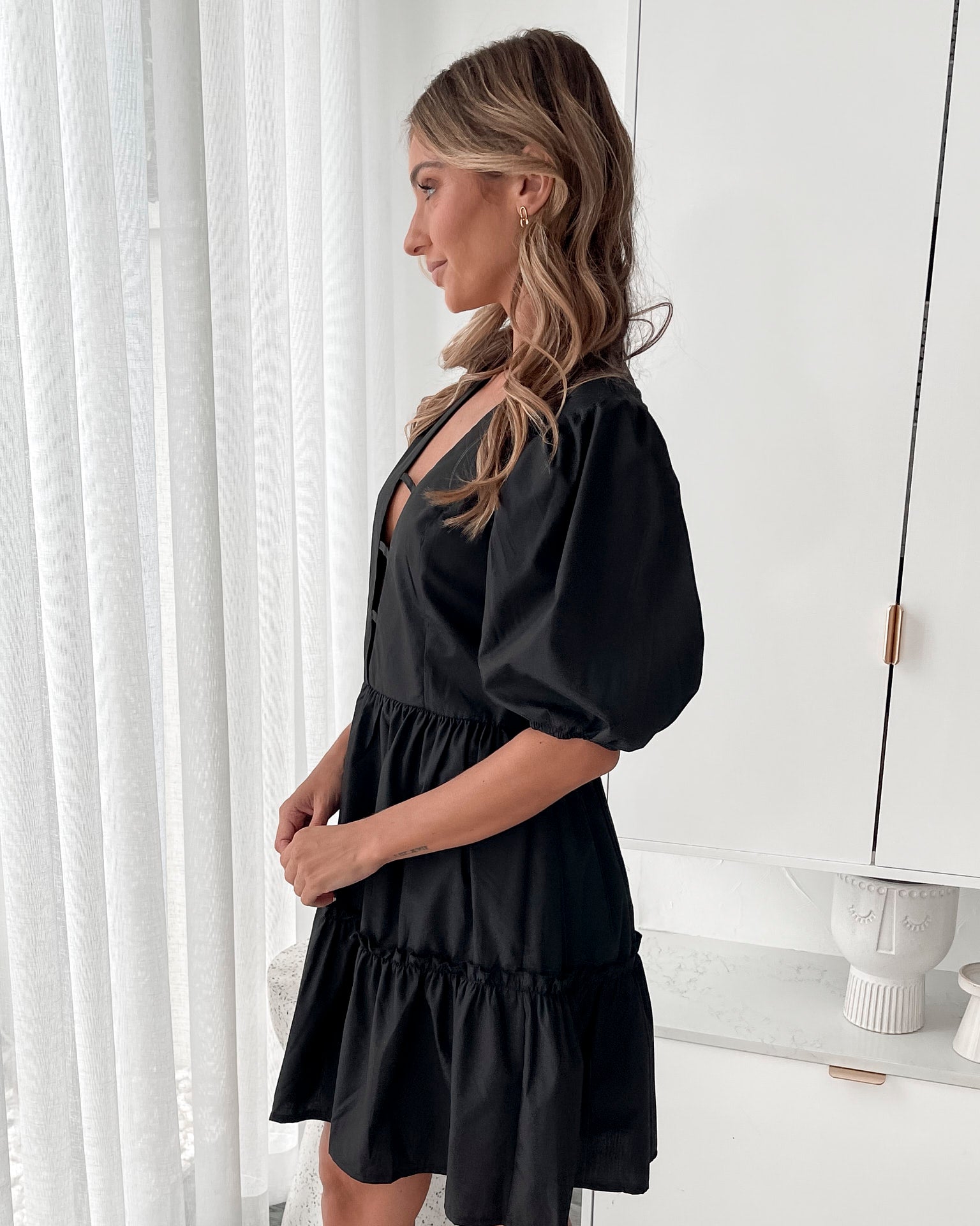 A woman wearing a classic black mini dress with a baby doll-inspired shape, featuring a plunge neckline and balloon sleeves.