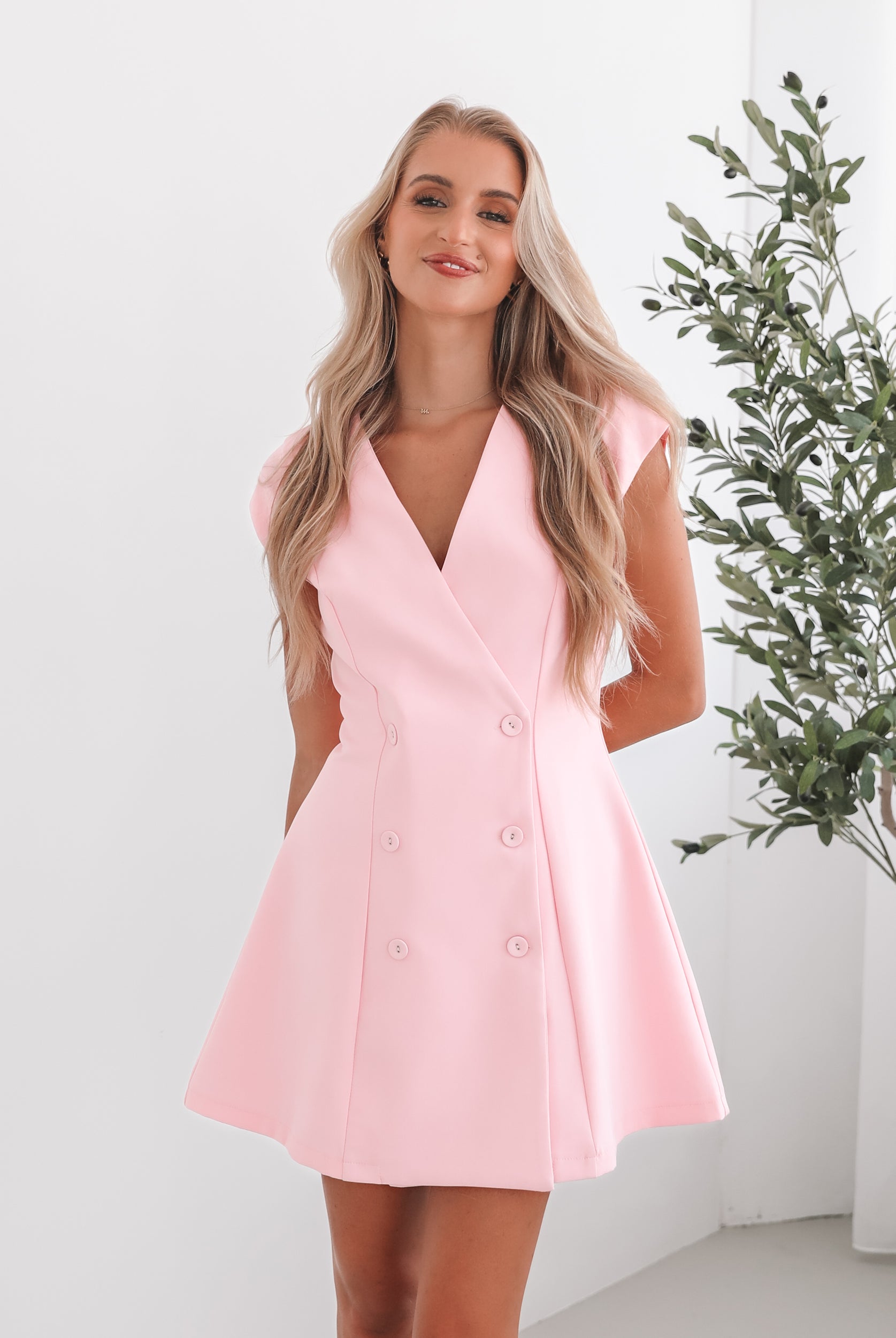 A woman wearing a pink mini dress with a v-neckline, cap sleeves, and buttons on the waist. The dress has a A-line silhouette.