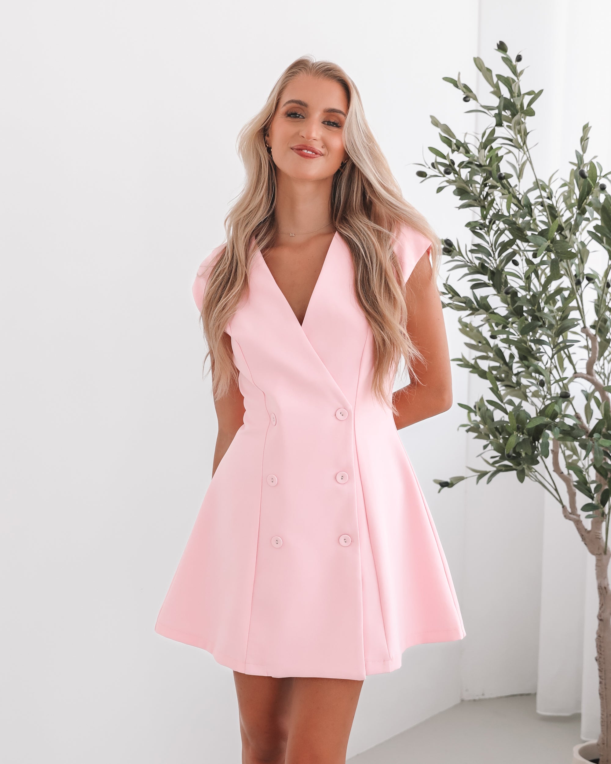 A woman wearing a pink mini dress with a v-neckline, cap sleeves, and buttons on the waist. The dress has a A-line silhouette.