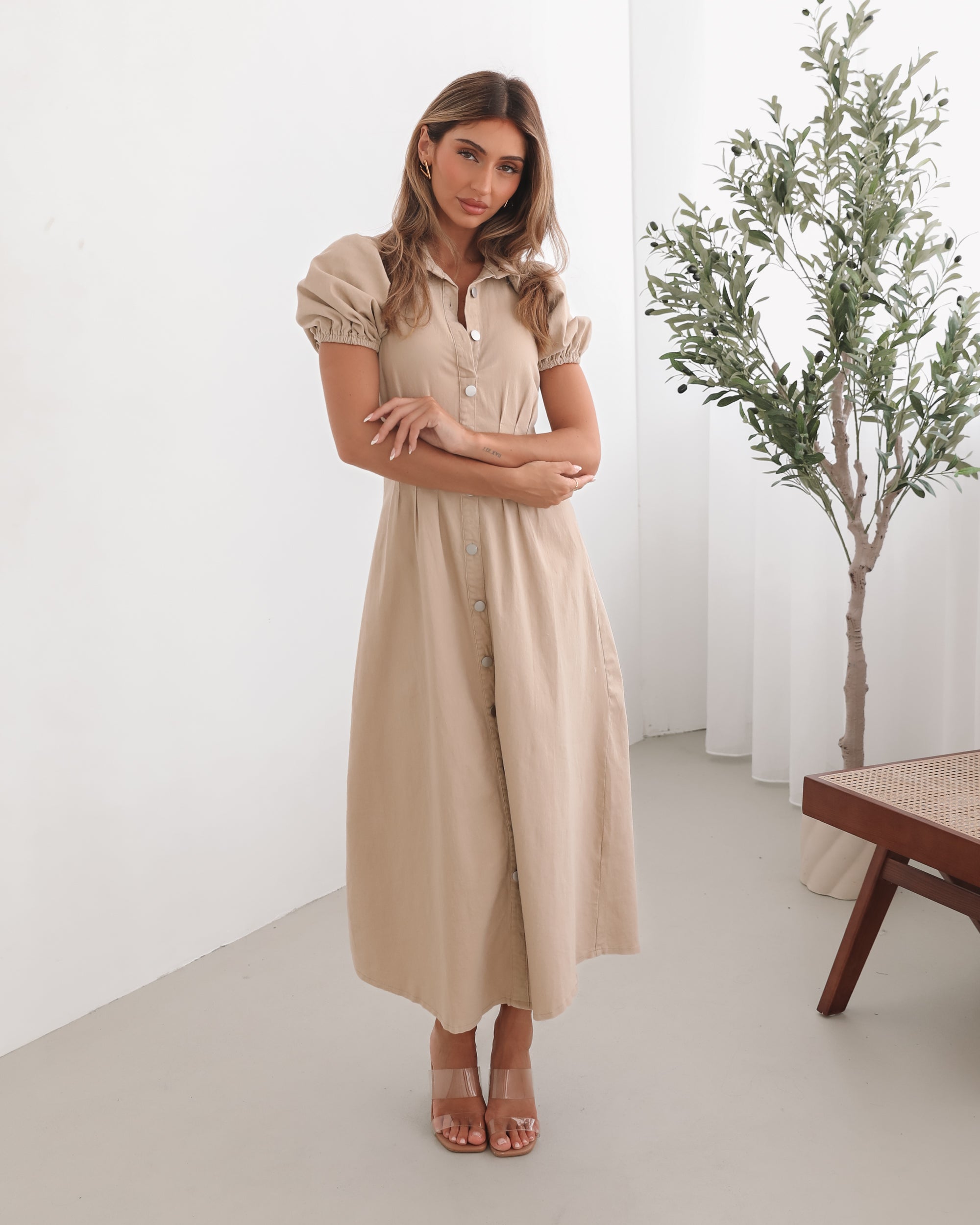 A woman standing in a room with a beige dress with a shirt collar, short puff sleeves, and a-line skirt with a midaxi length hem.