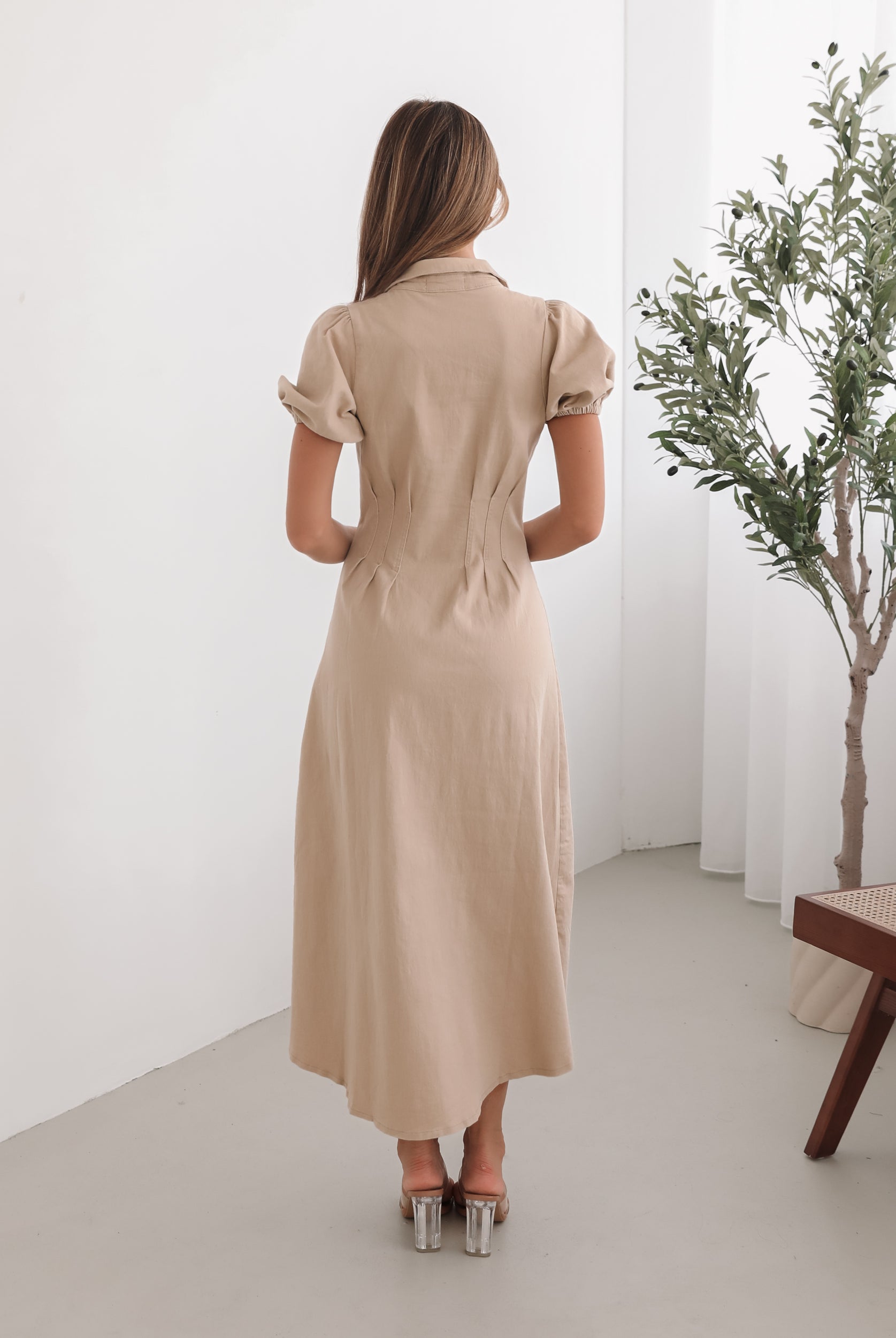 A woman standing in a room with a beige dress with a shirt collar, short puff sleeves, and a-line skirt with a midaxi length hem.