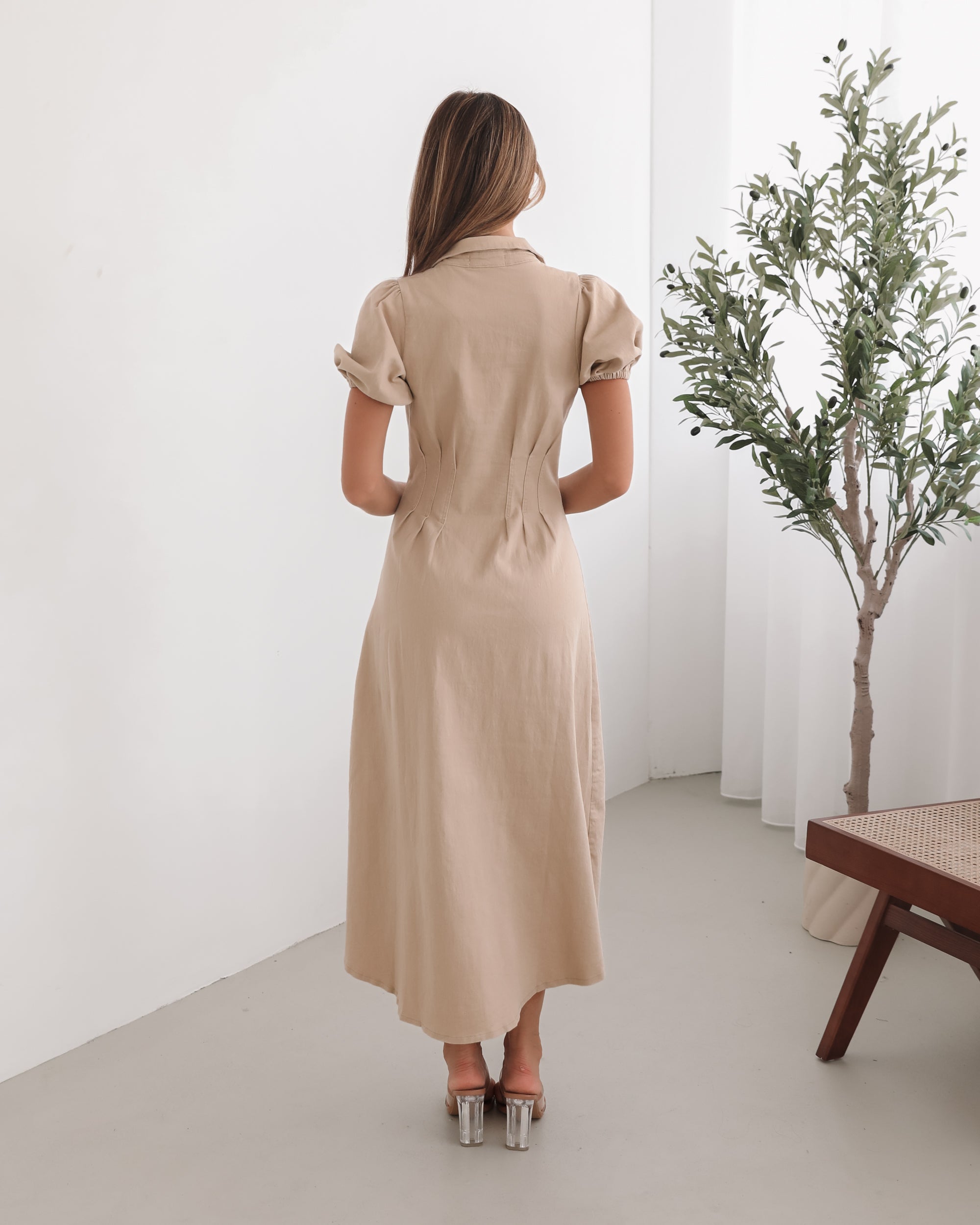 A woman standing in a room with a beige dress with a shirt collar, short puff sleeves, and a-line skirt with a midaxi length hem.