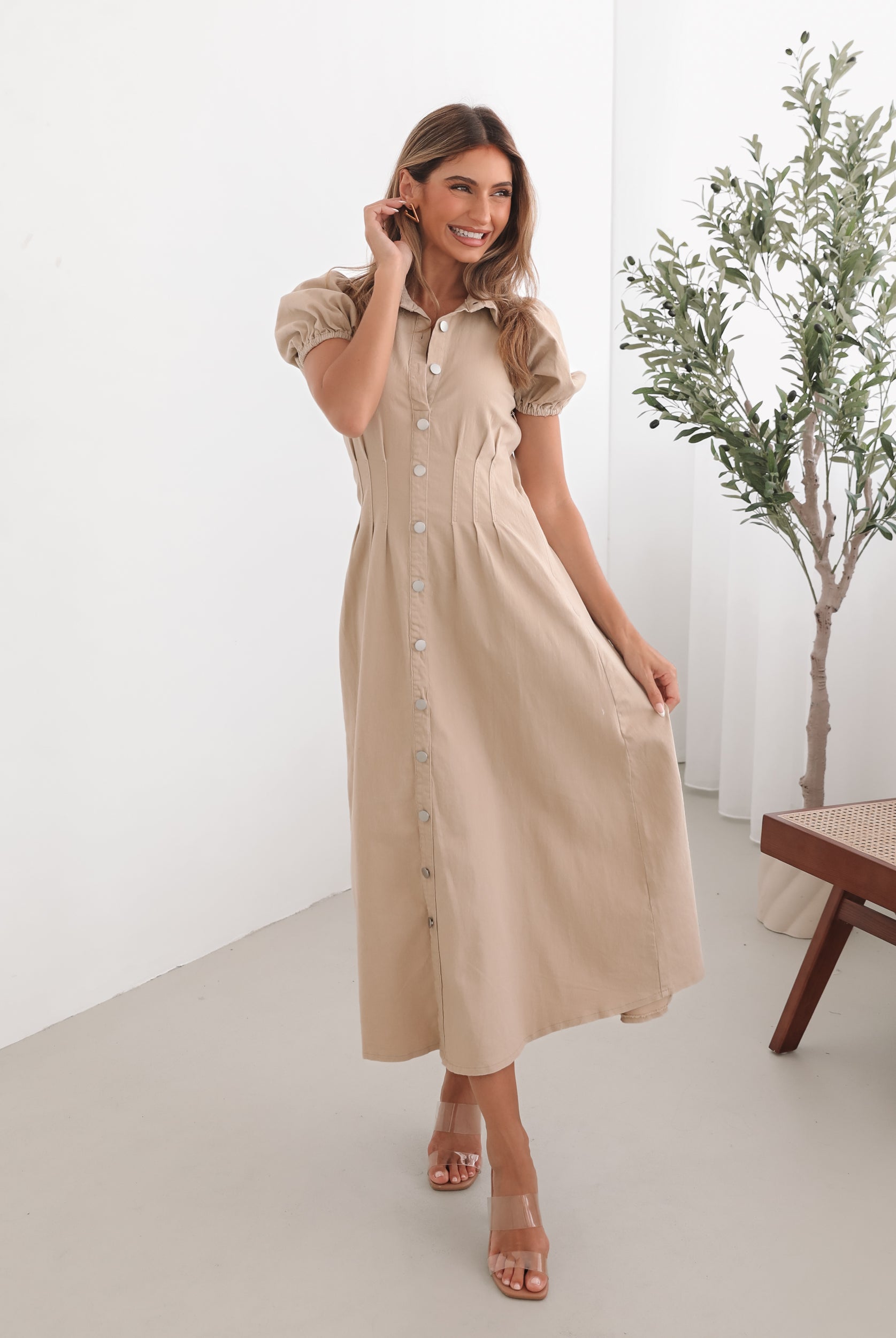 A woman standing in a room with a beige dress with a shirt collar, short puff sleeves, and a-line skirt with a midaxi length hem.