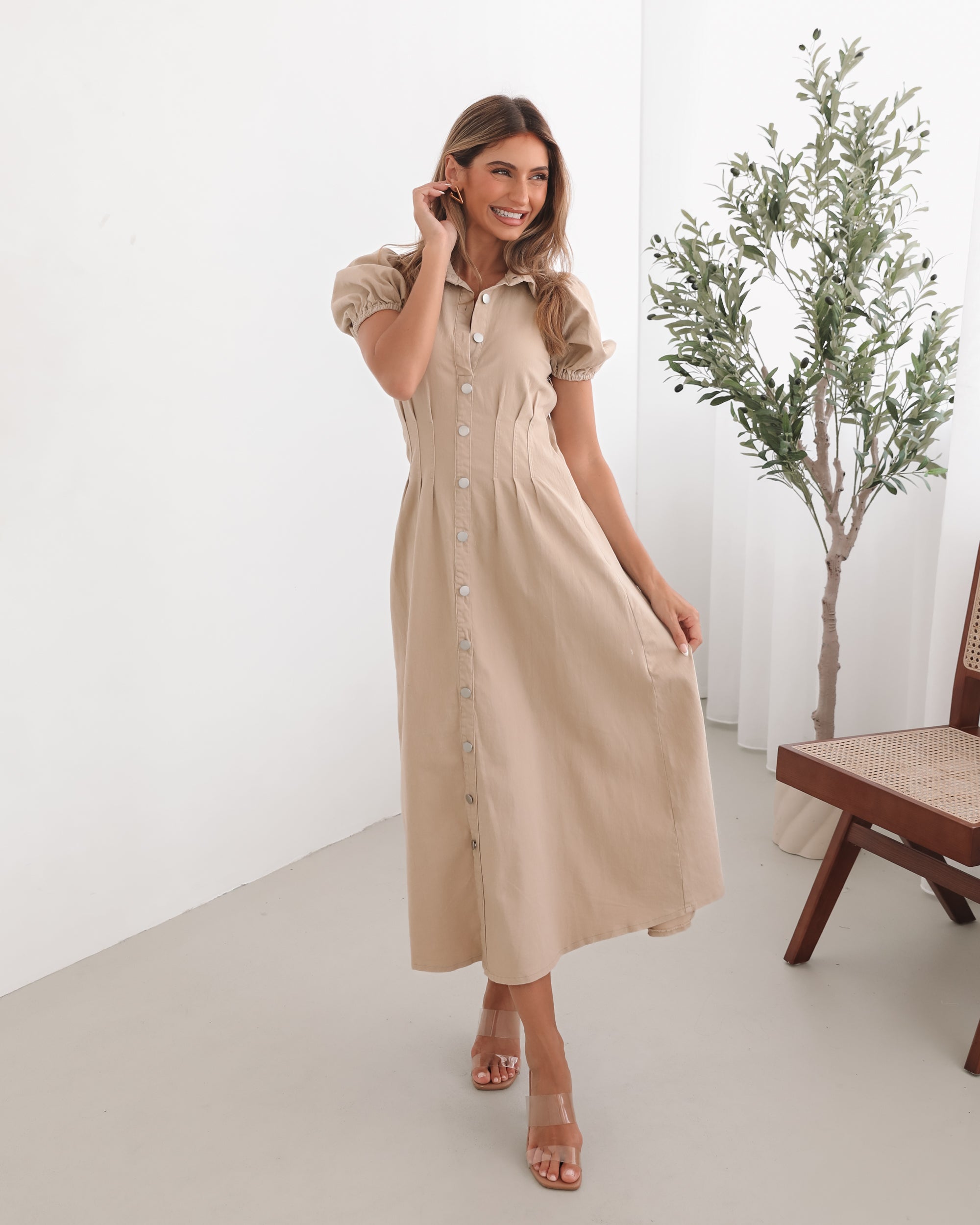 A woman standing in a room with a beige dress with a shirt collar, short puff sleeves, and a-line skirt with a midaxi length hem.