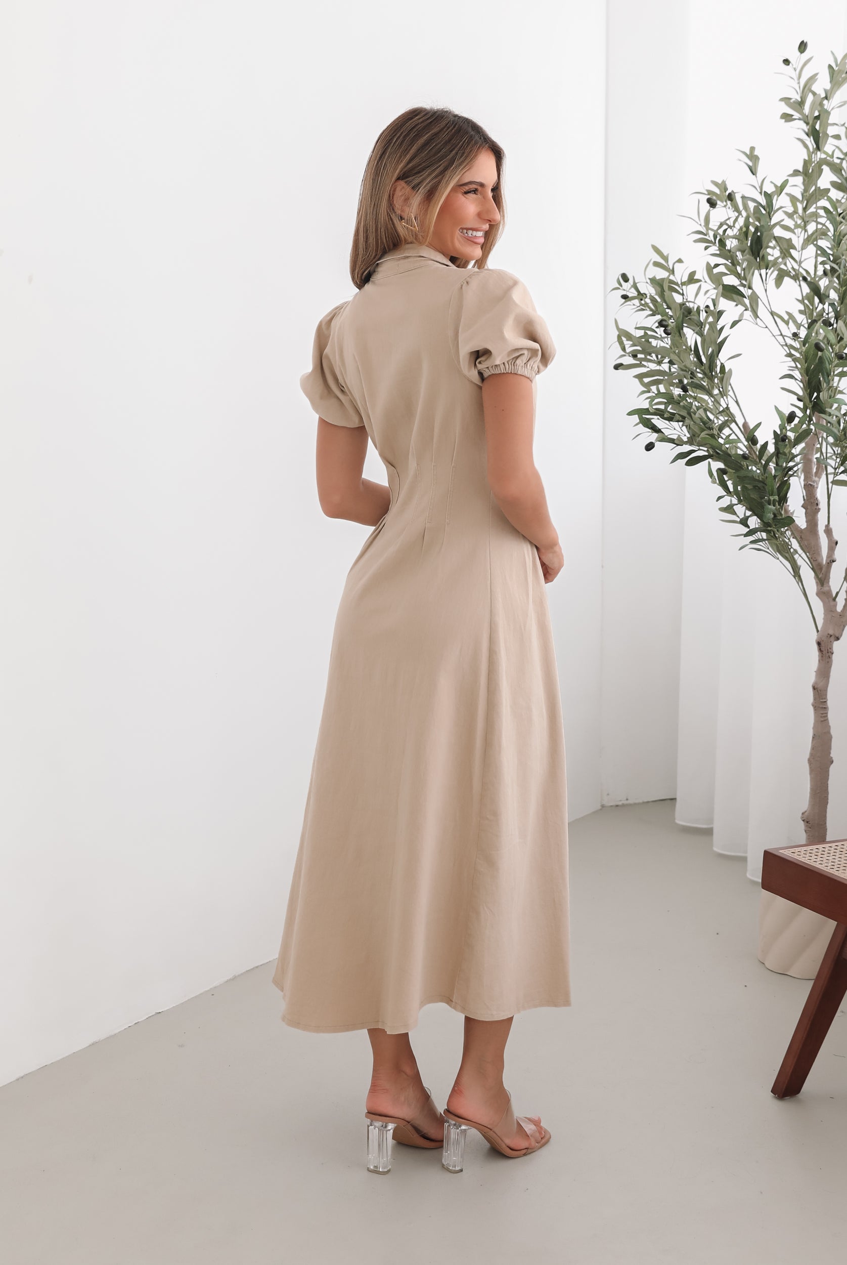 A woman standing in a room with a beige dress with a shirt collar, short puff sleeves, and a-line skirt with a midaxi length hem.
