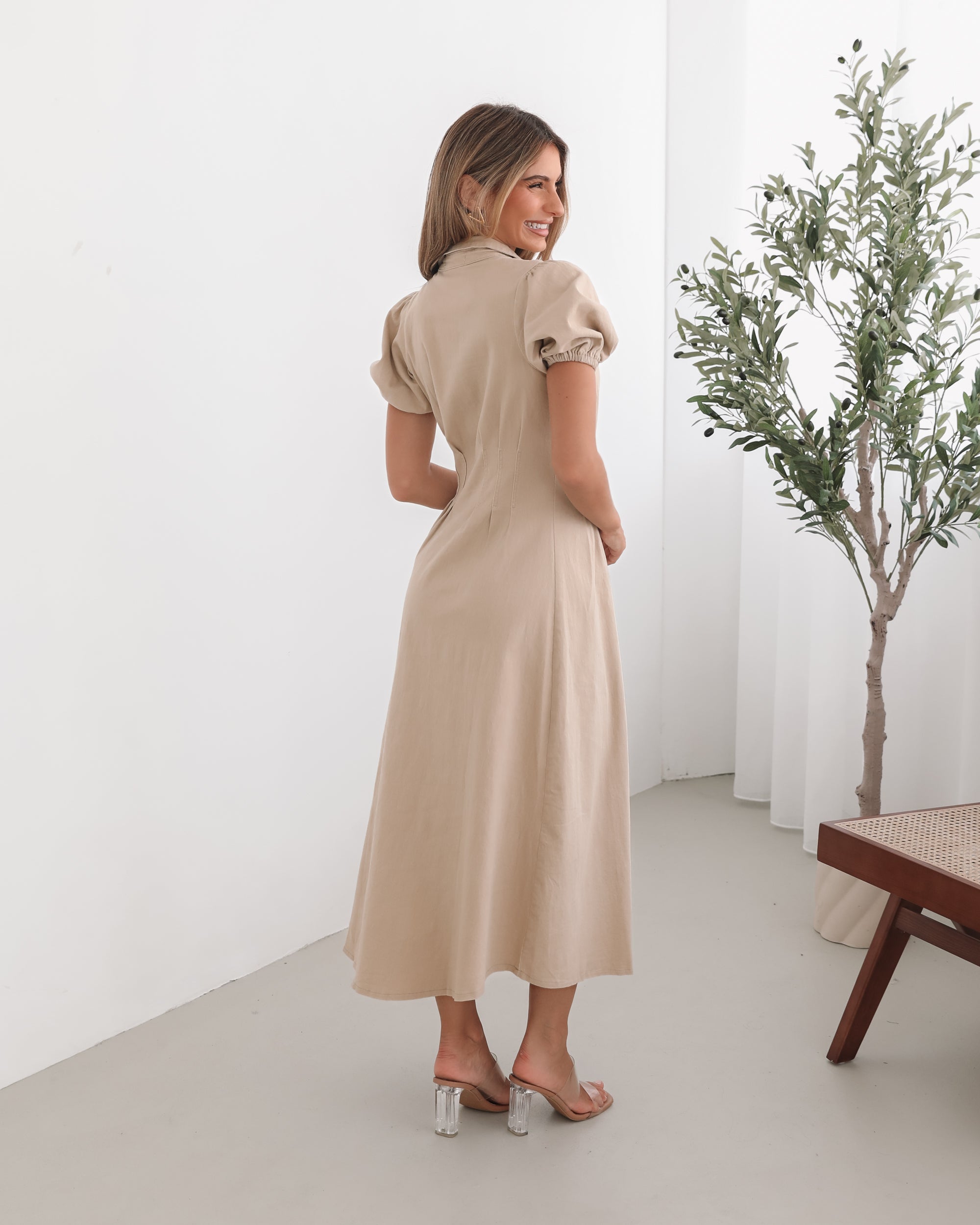 A woman standing in a room with a beige dress with a shirt collar, short puff sleeves, and a-line skirt with a midaxi length hem.