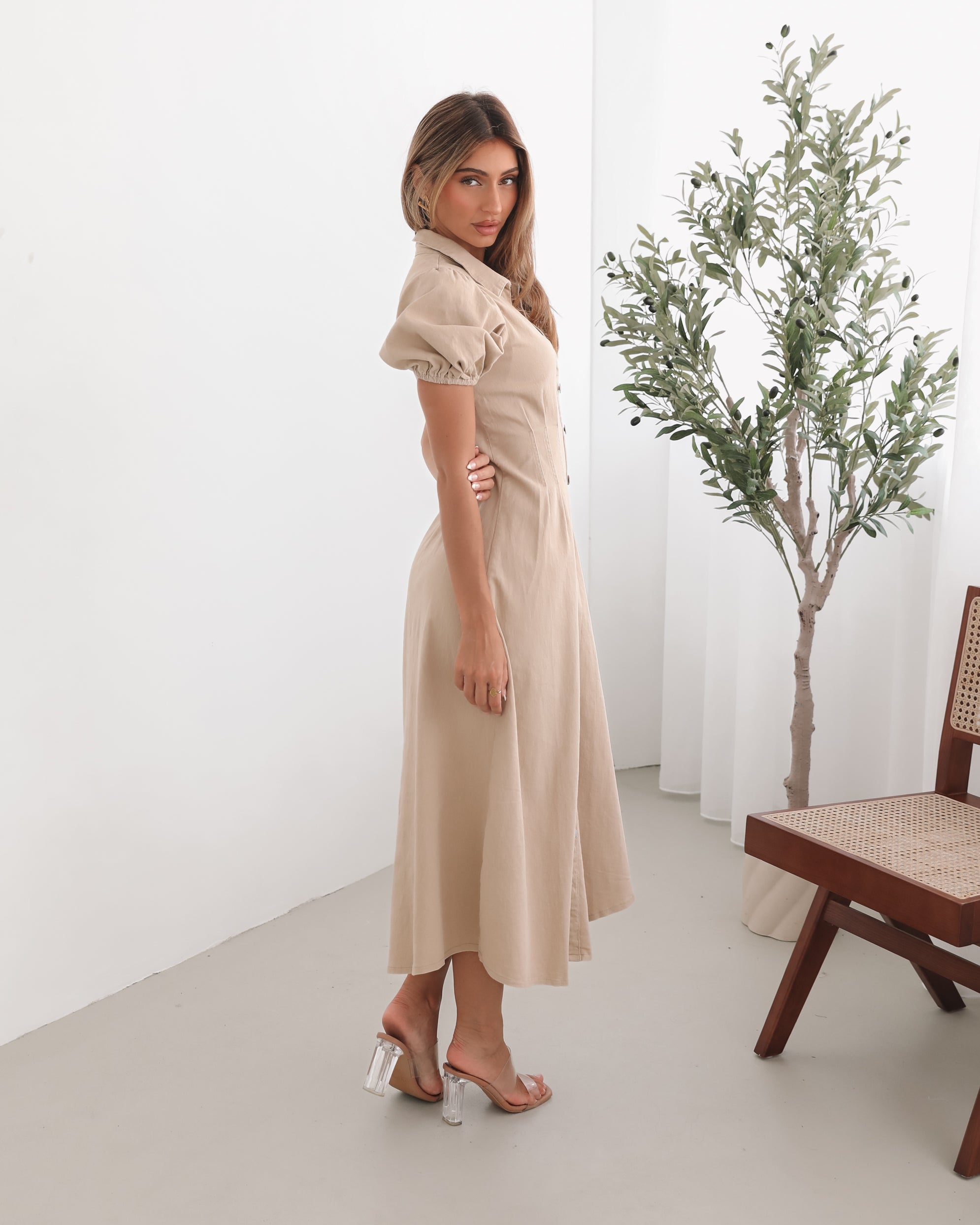 A woman standing in a room with a beige dress with a shirt collar, short puff sleeves, and a-line skirt with a midaxi length hem.