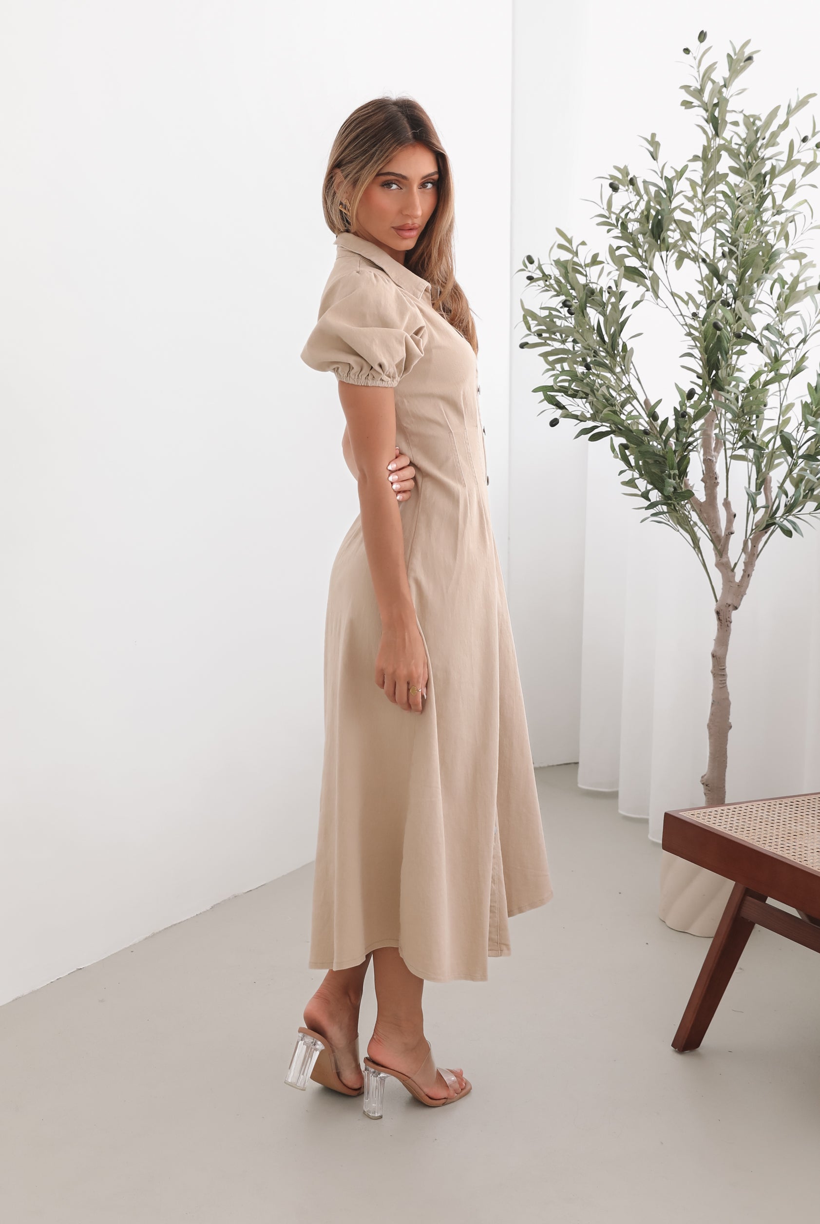 A woman standing in a room with a beige dress with a shirt collar, short puff sleeves, and a-line skirt with a midaxi length hem.