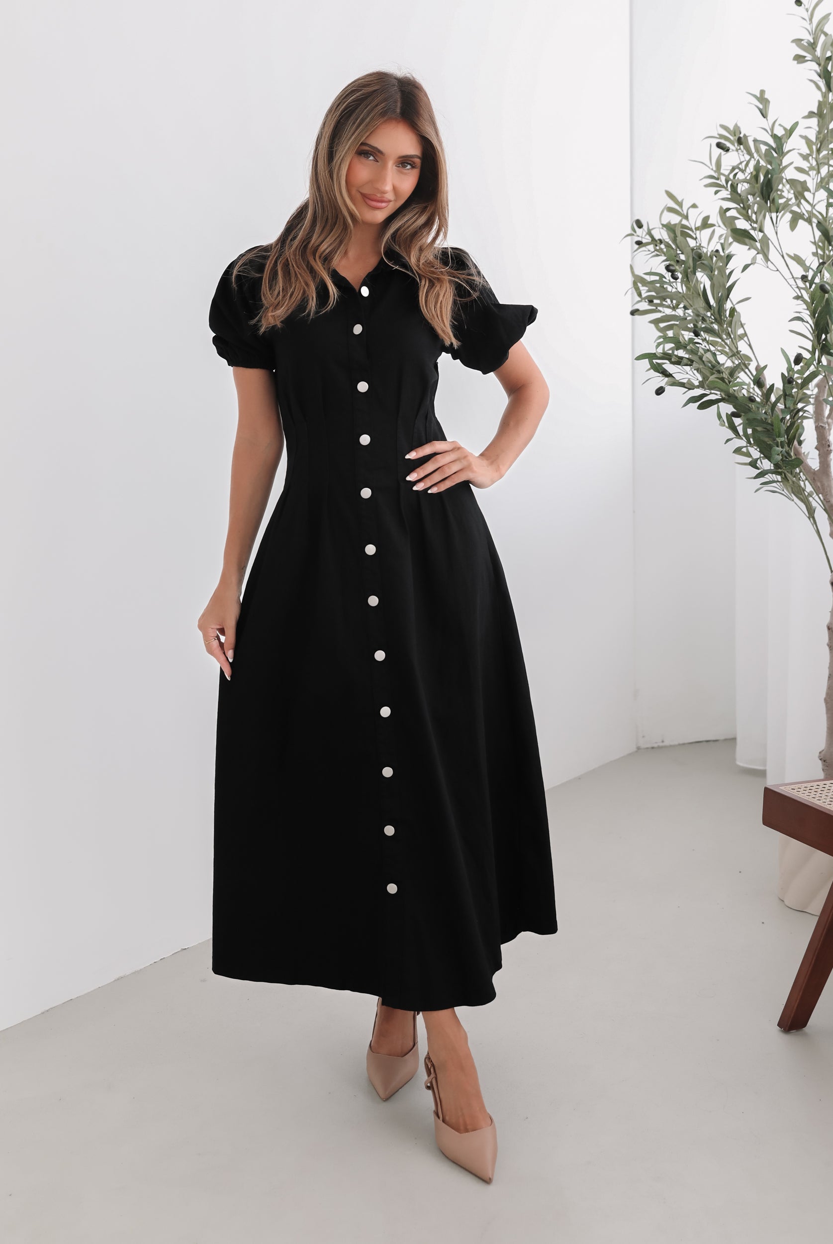 A woman standing in a room with a white background wearing a black knee-length dress with a shirt collar, short puff sleeves, and center-front buttons.
