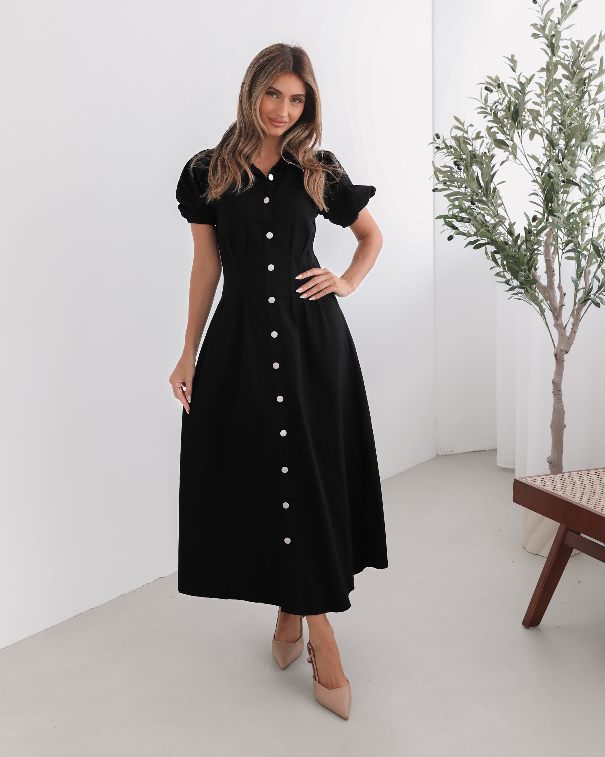 A woman standing in a room with a white background wearing a black knee-length dress with a shirt collar, short puff sleeves, and center-front buttons.