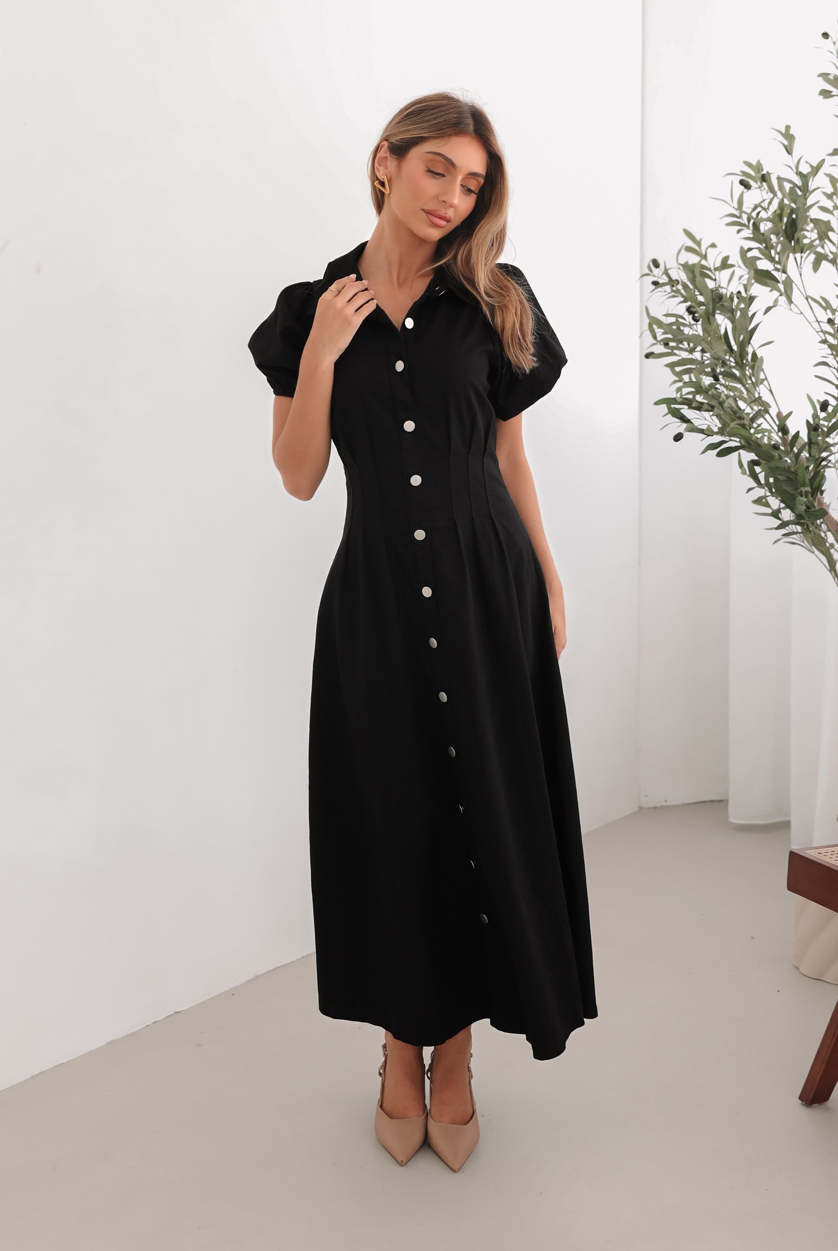 A woman standing in a room with a white background wearing a black knee-length dress with a shirt collar, short puff sleeves, and center-front buttons.