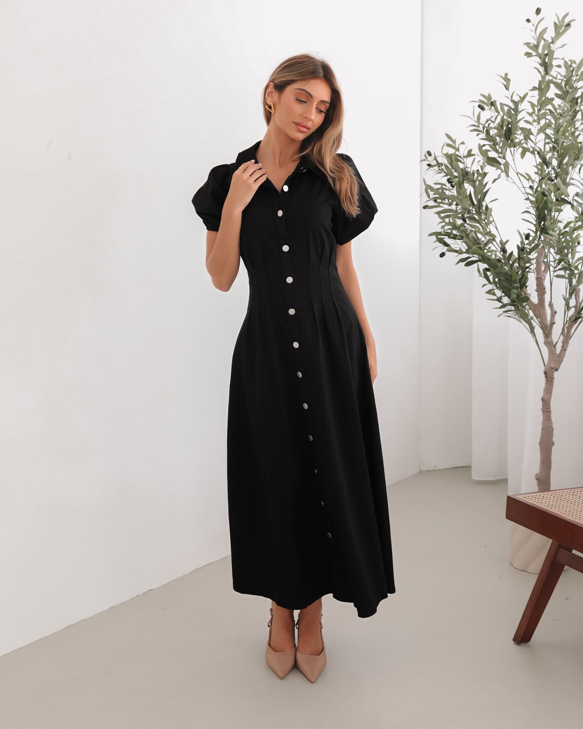 A woman standing in a room with a white background wearing a black knee-length dress with a shirt collar, short puff sleeves, and center-front buttons.