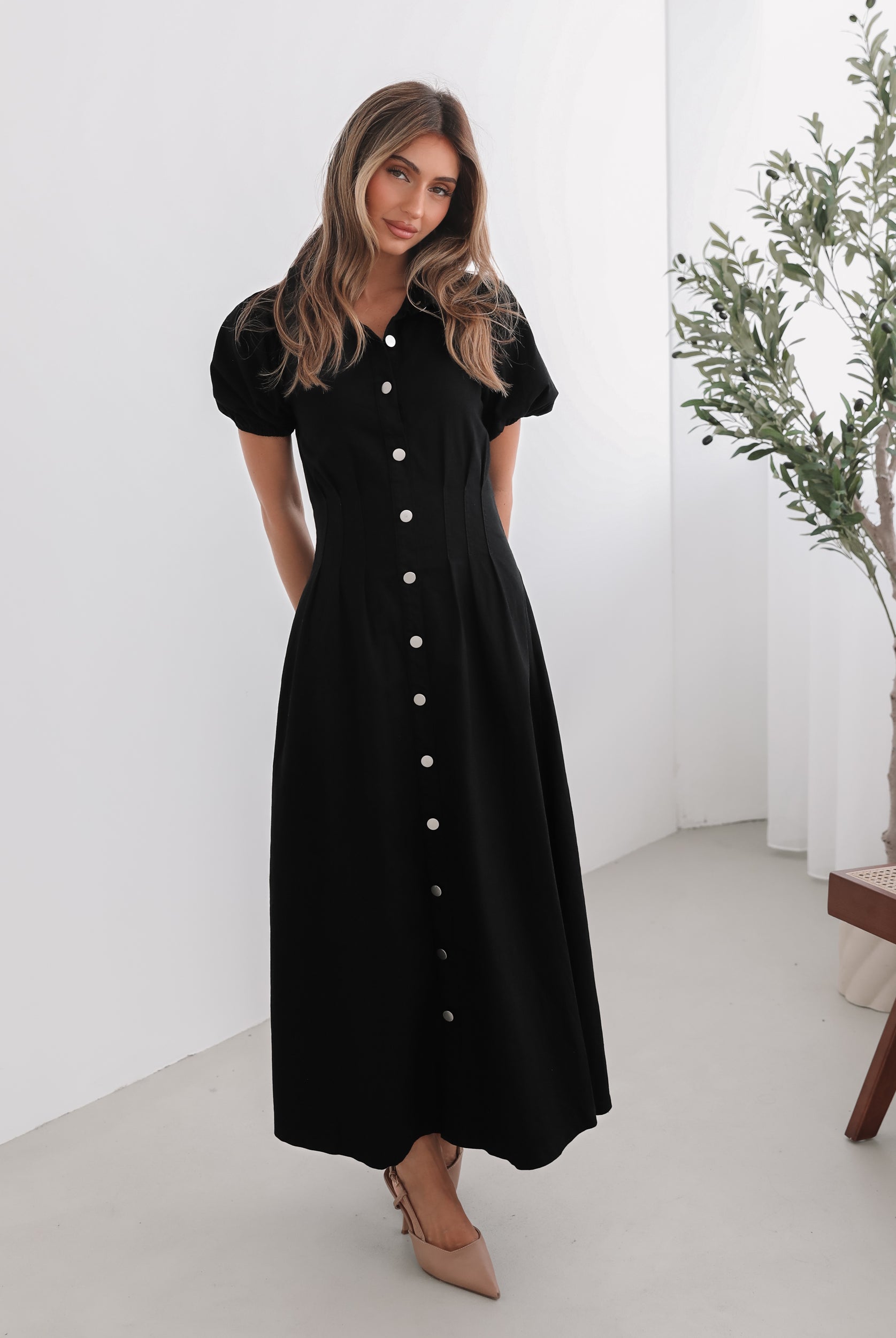 A woman standing in a room with a white background wearing a black knee-length dress with a shirt collar, short puff sleeves, and center-front buttons.