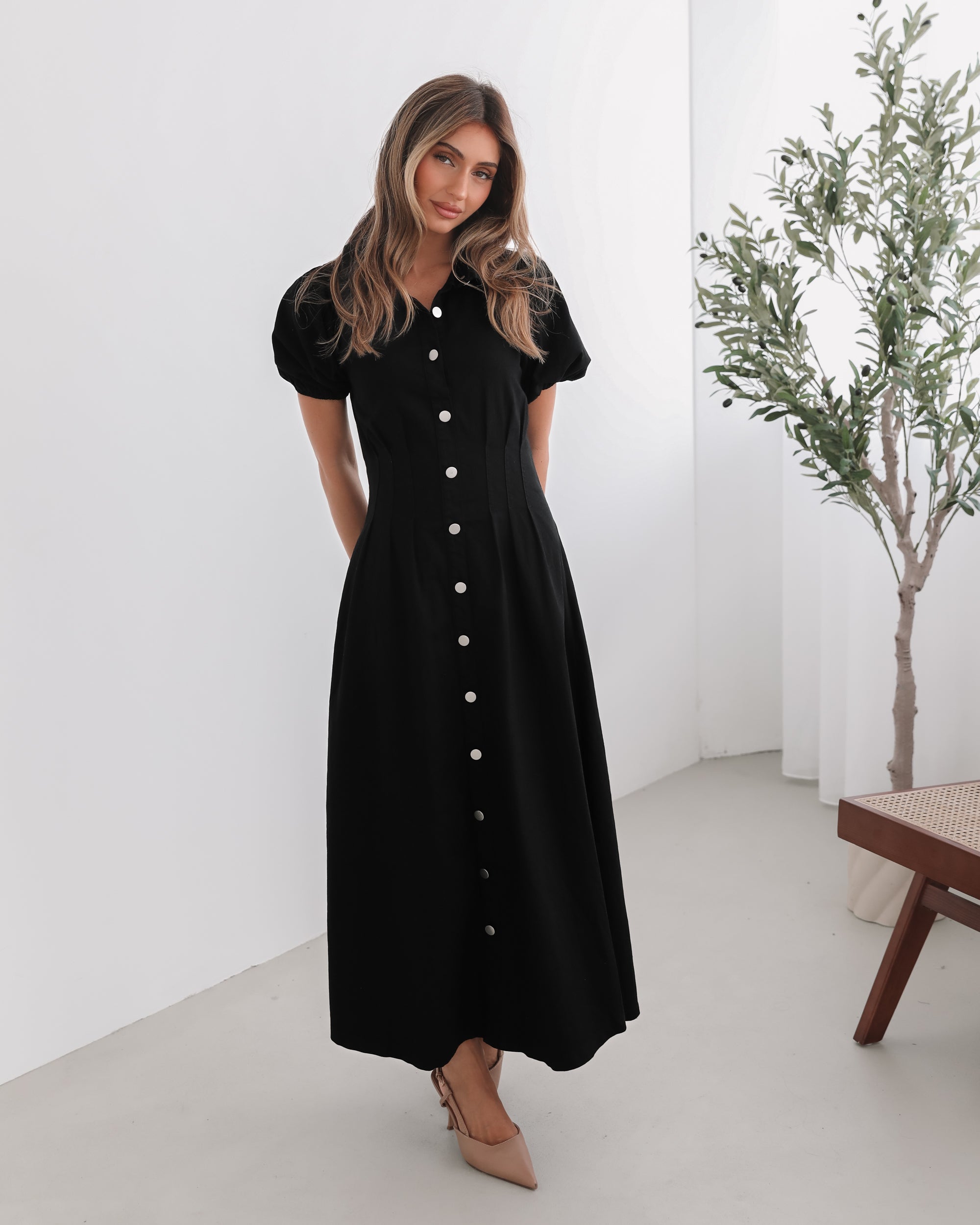 A woman standing in a room with a white background wearing a black knee-length dress with a shirt collar, short puff sleeves, and center-front buttons.